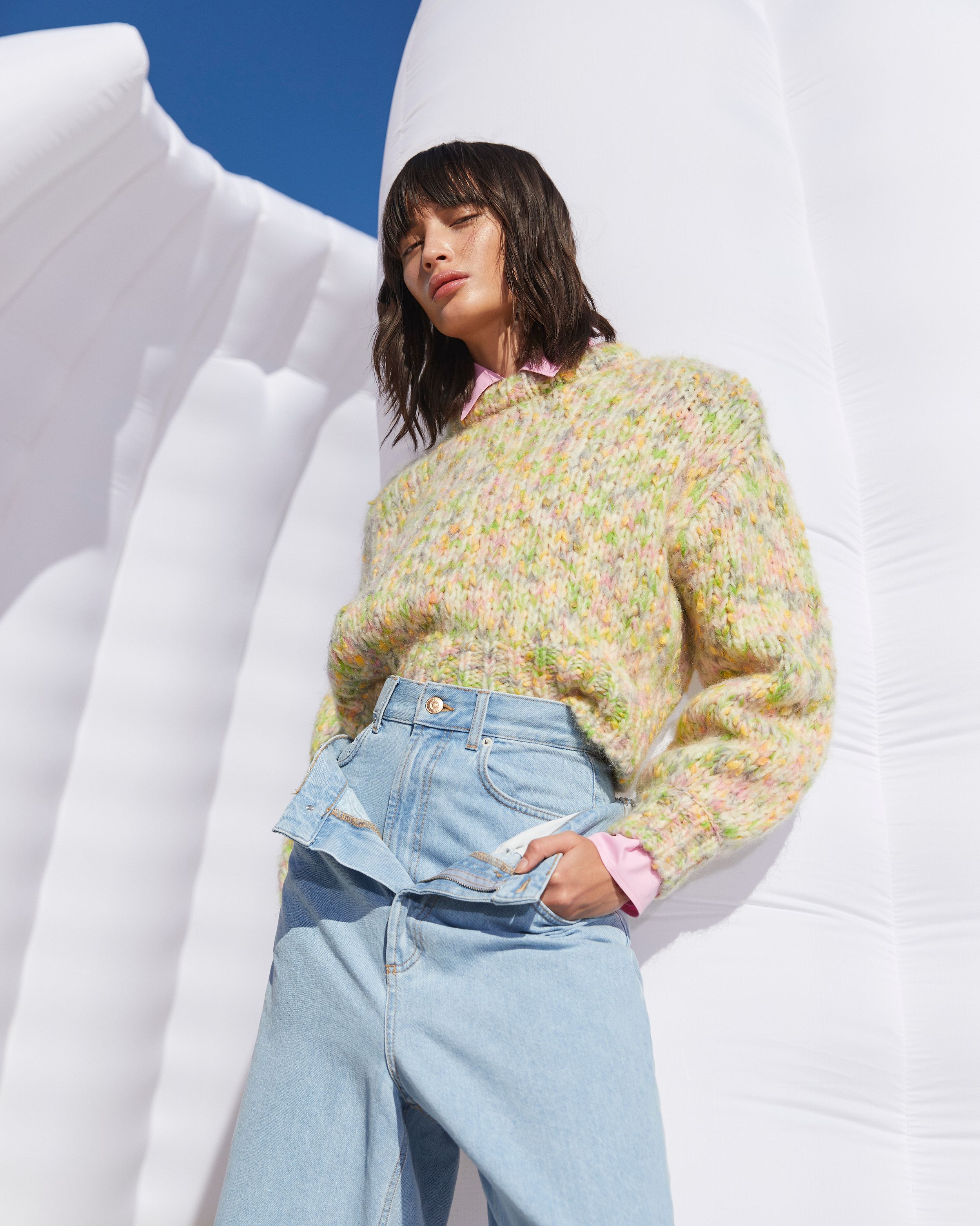 Model standing in front of white inflatable structure wearing a pastel multi-coloured knit and bleached jeans