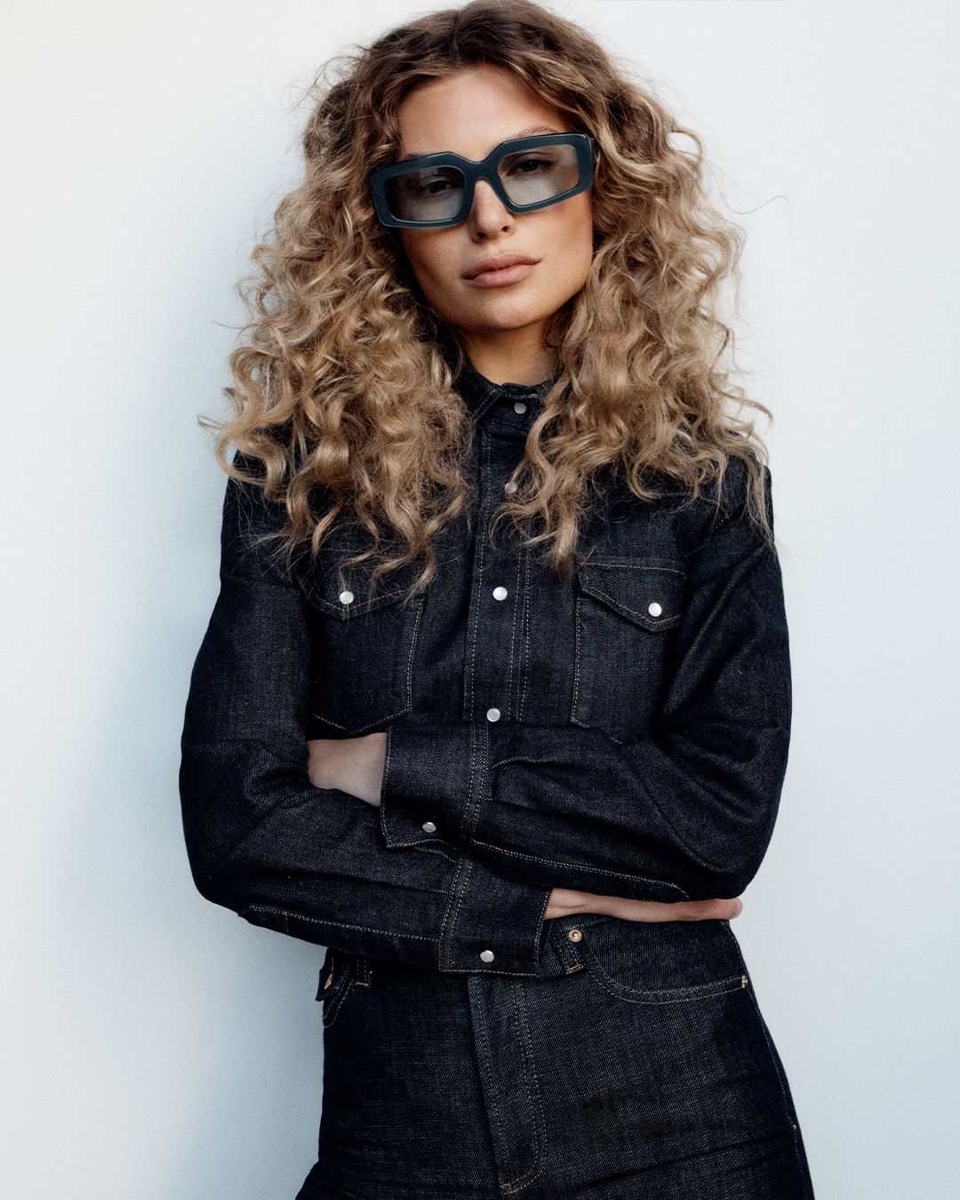Model standing with arms crossed wearing blue sunglasses with dark blue denim shirt and jeans