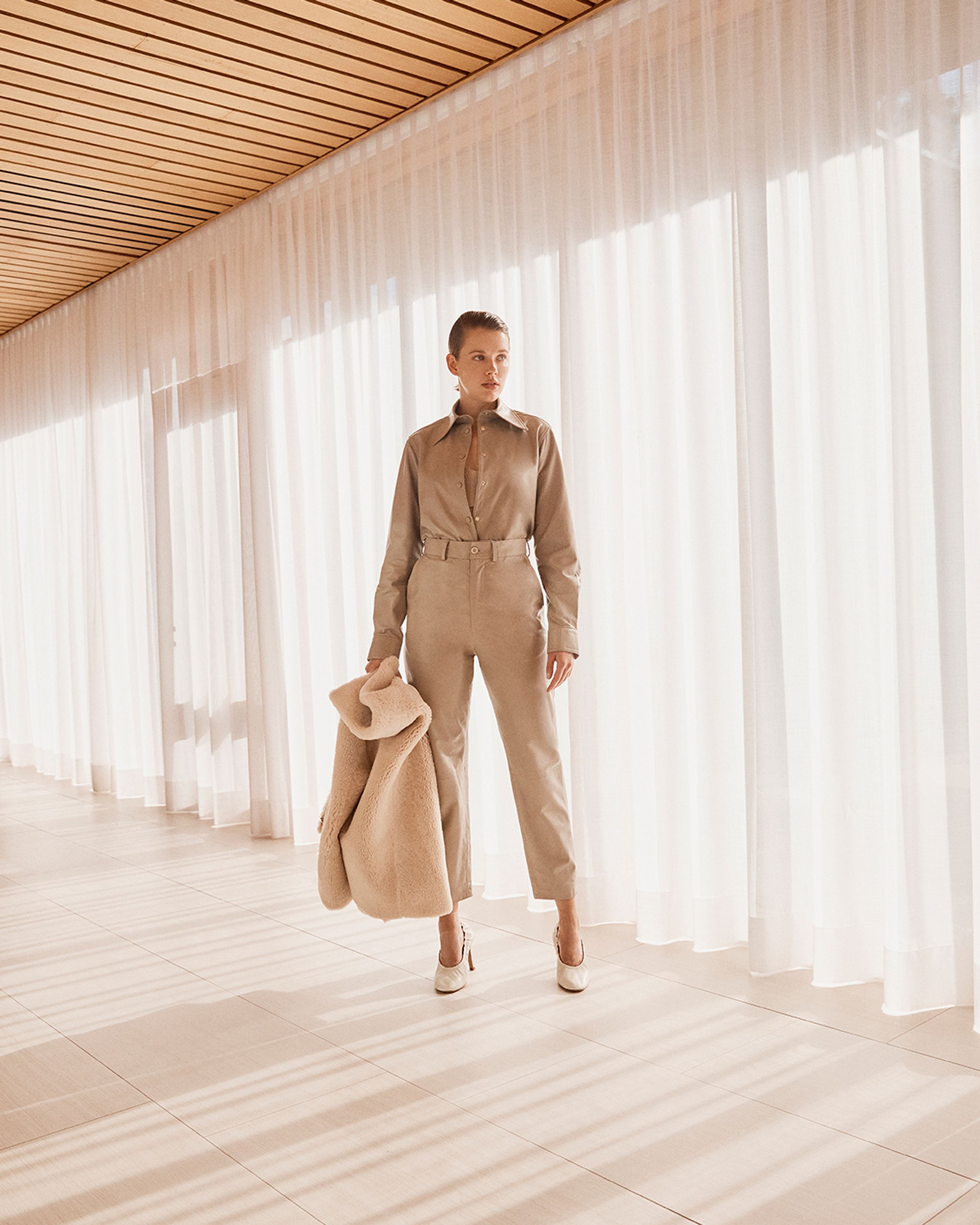 Model standing by a window wearing neutral leather shirt and trousers, holding a shearling jacket