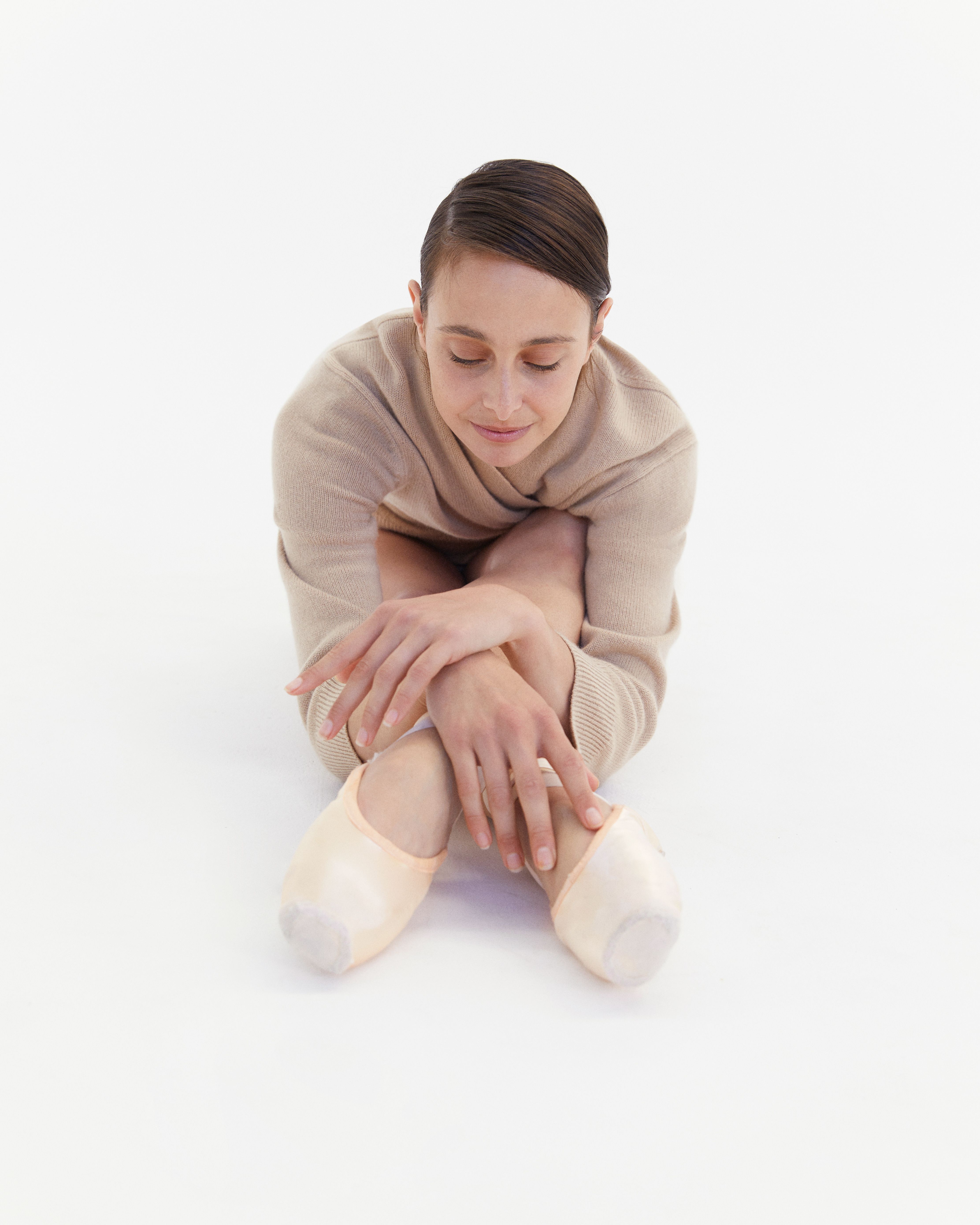 Dimity Azoury sitting down leaning over her feet wearing a beige knit and pointe shoes