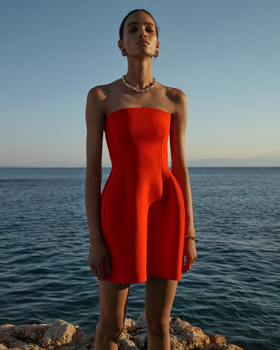 Model standing in front of the ocean wearing a orange strapless mini dress