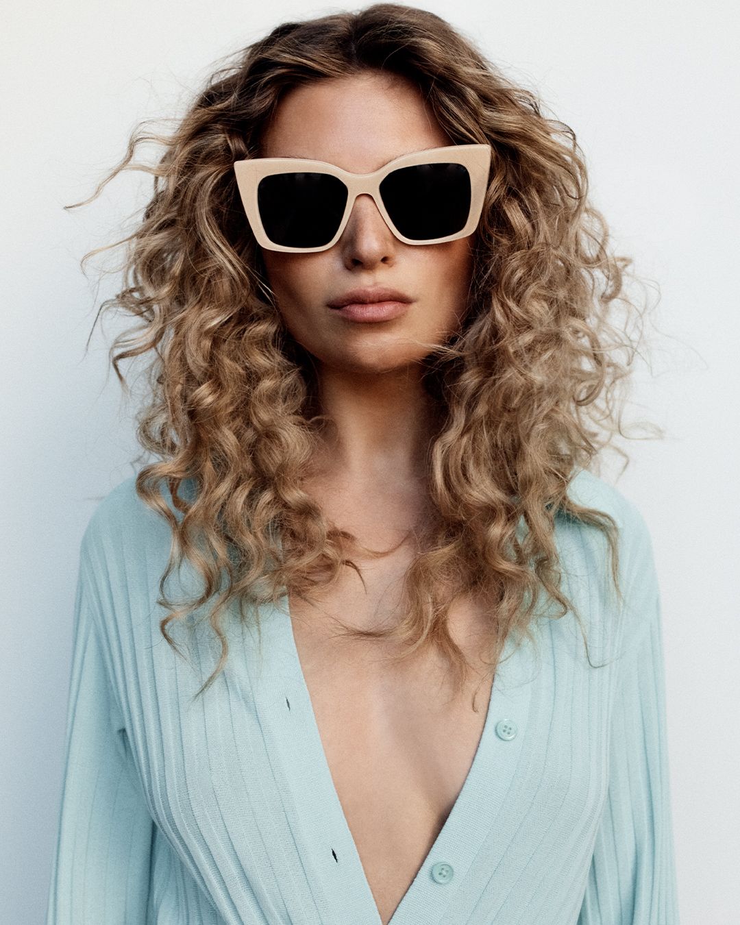 Model facing the camera wearing light beige oversized sunglasses and a light blue knitted top