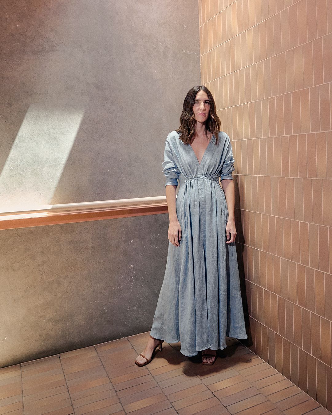 Bianca Marchi wearing a blue grey linen maxi dress standing in a brown tile and concrete hallway