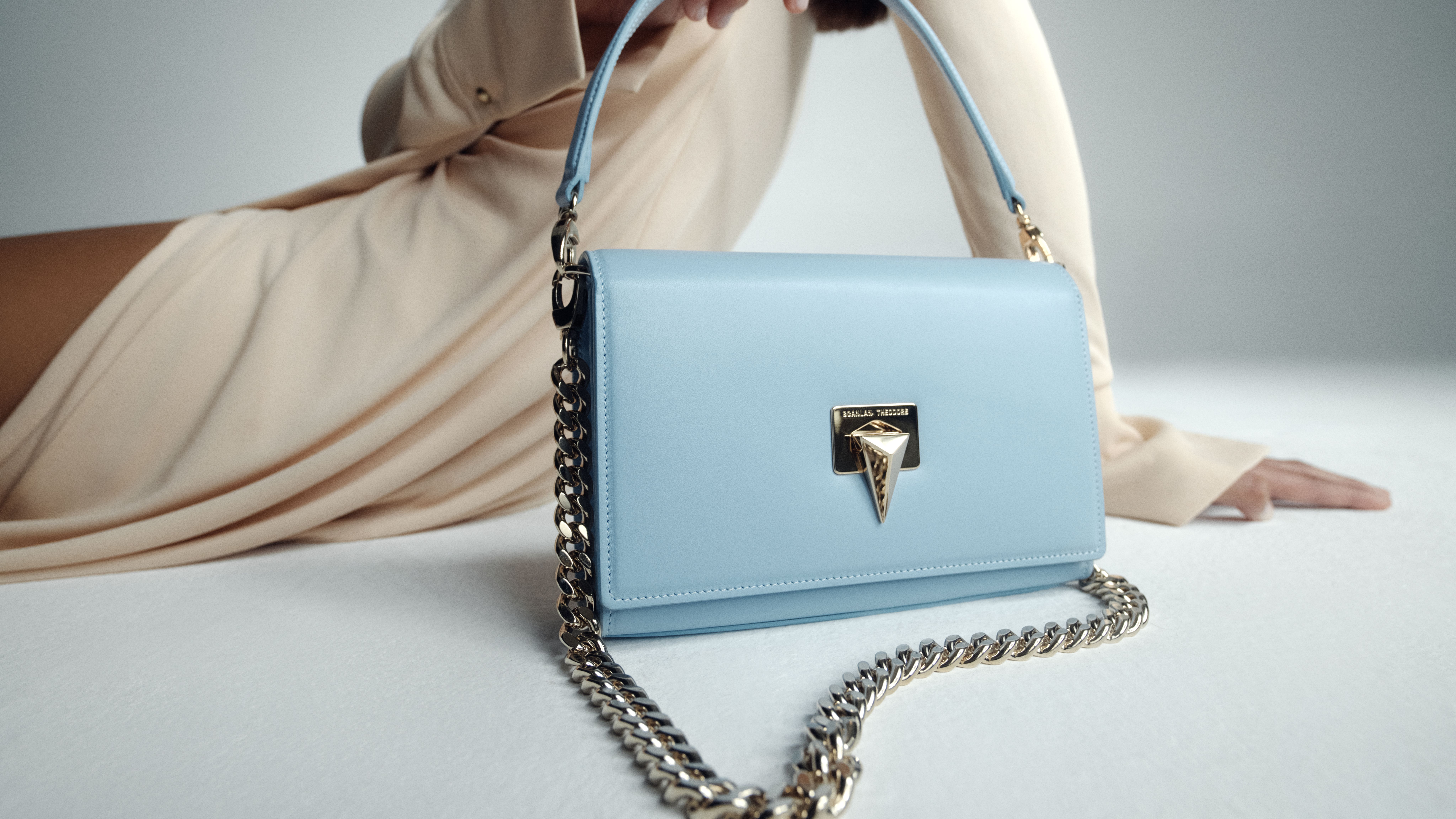 Close up image of pale blue triangle chain bag with model lying behind in a sand coloured long sleeve dress