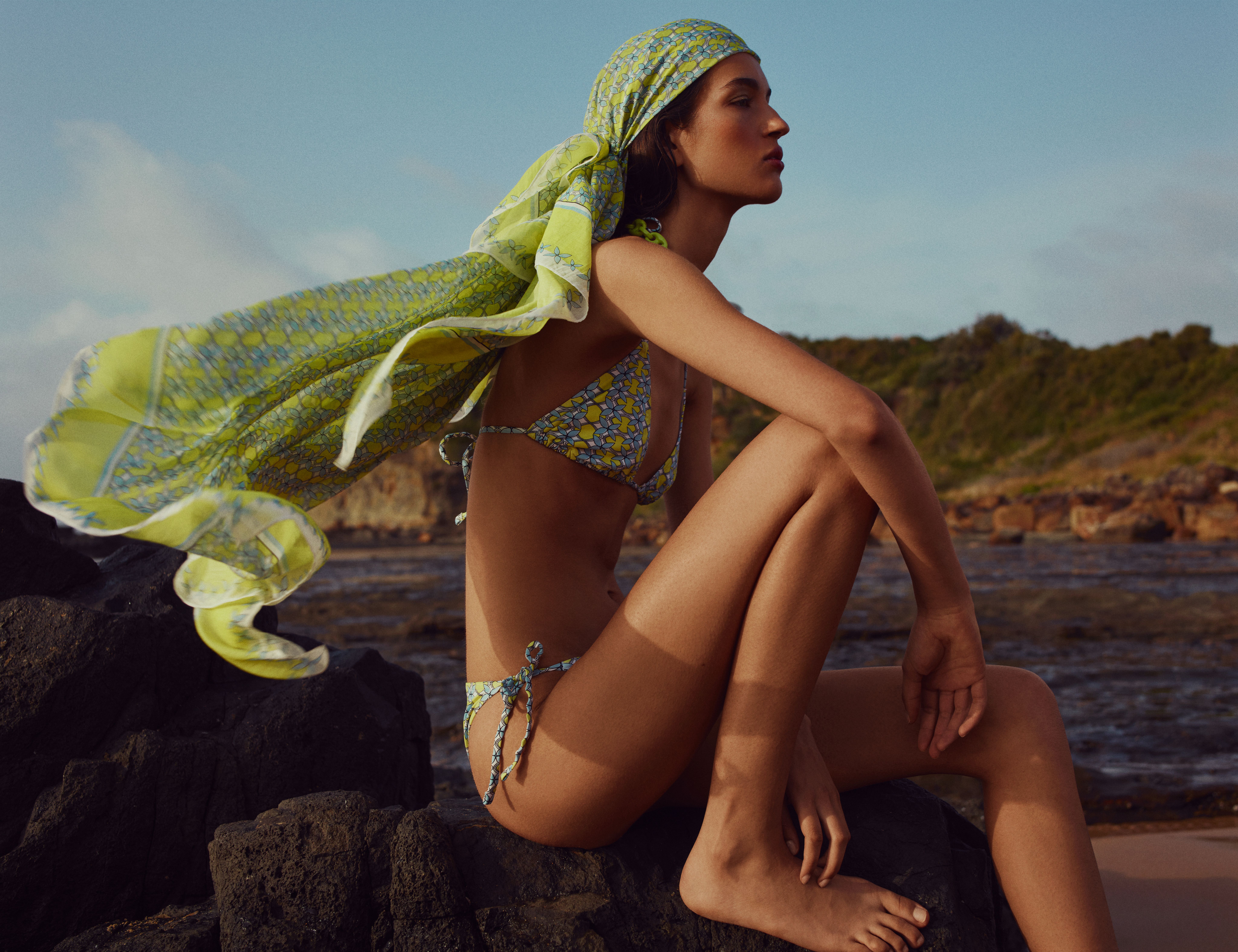 Model sitting facing the side wearing a green and blue bikini and scarf sitting on rocks with the ocean in the background