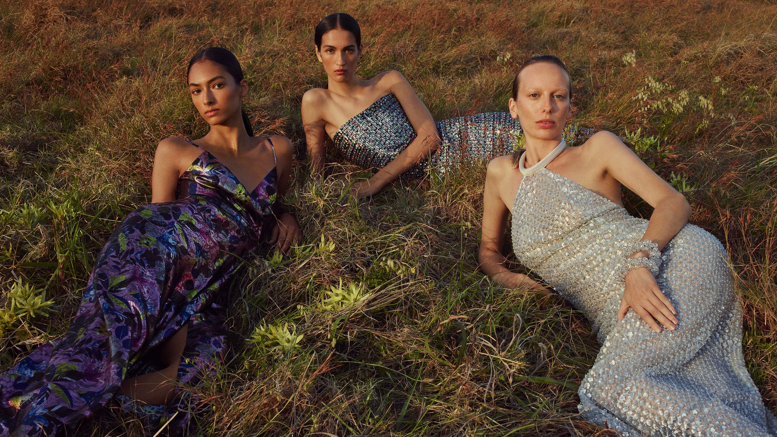 Three models lying on the grass wearing violet, silver and multicoloured evening gowns