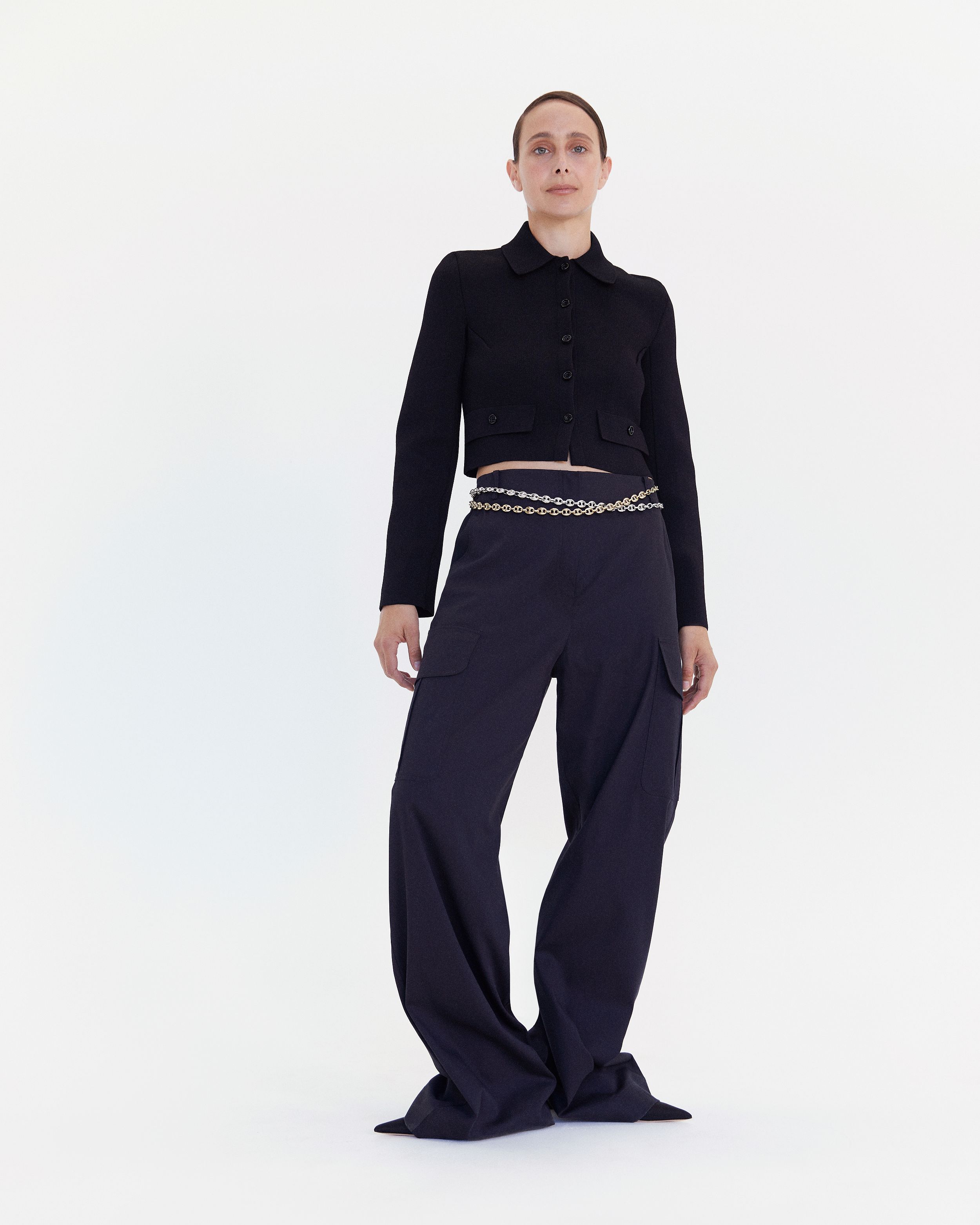 Dimity Azoury standing tall wearing a black cropped jacket and long navy trousers with silver and gold chain belts