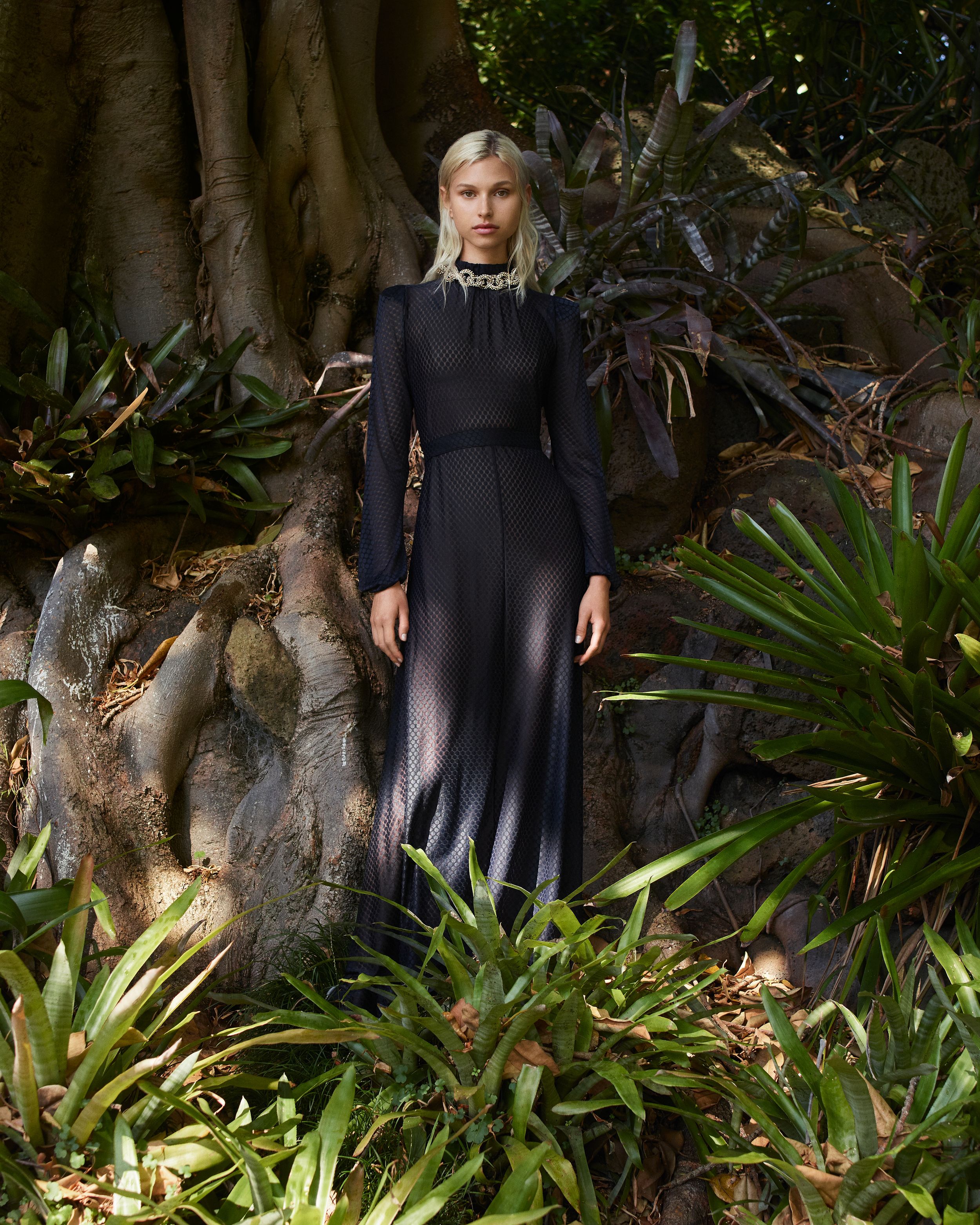 Model standing in front of a fig tree wearing a black sheer long-sleeved gown with chunky gold necklace