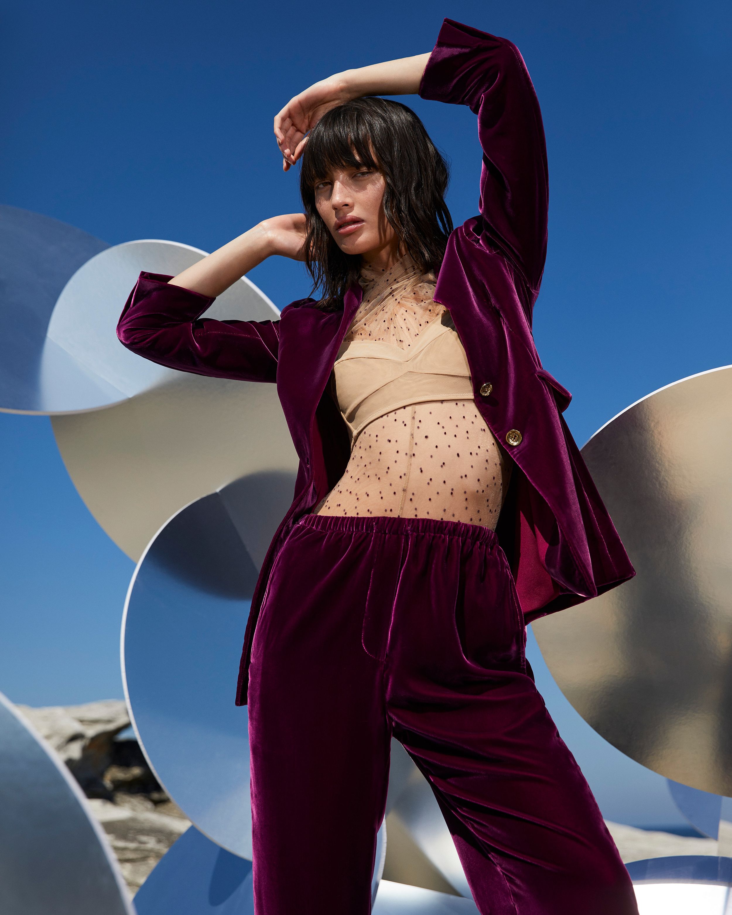 Model standing amongst mirrored sculptures outdoors wearing a mulberry velvet suit and beige blouse