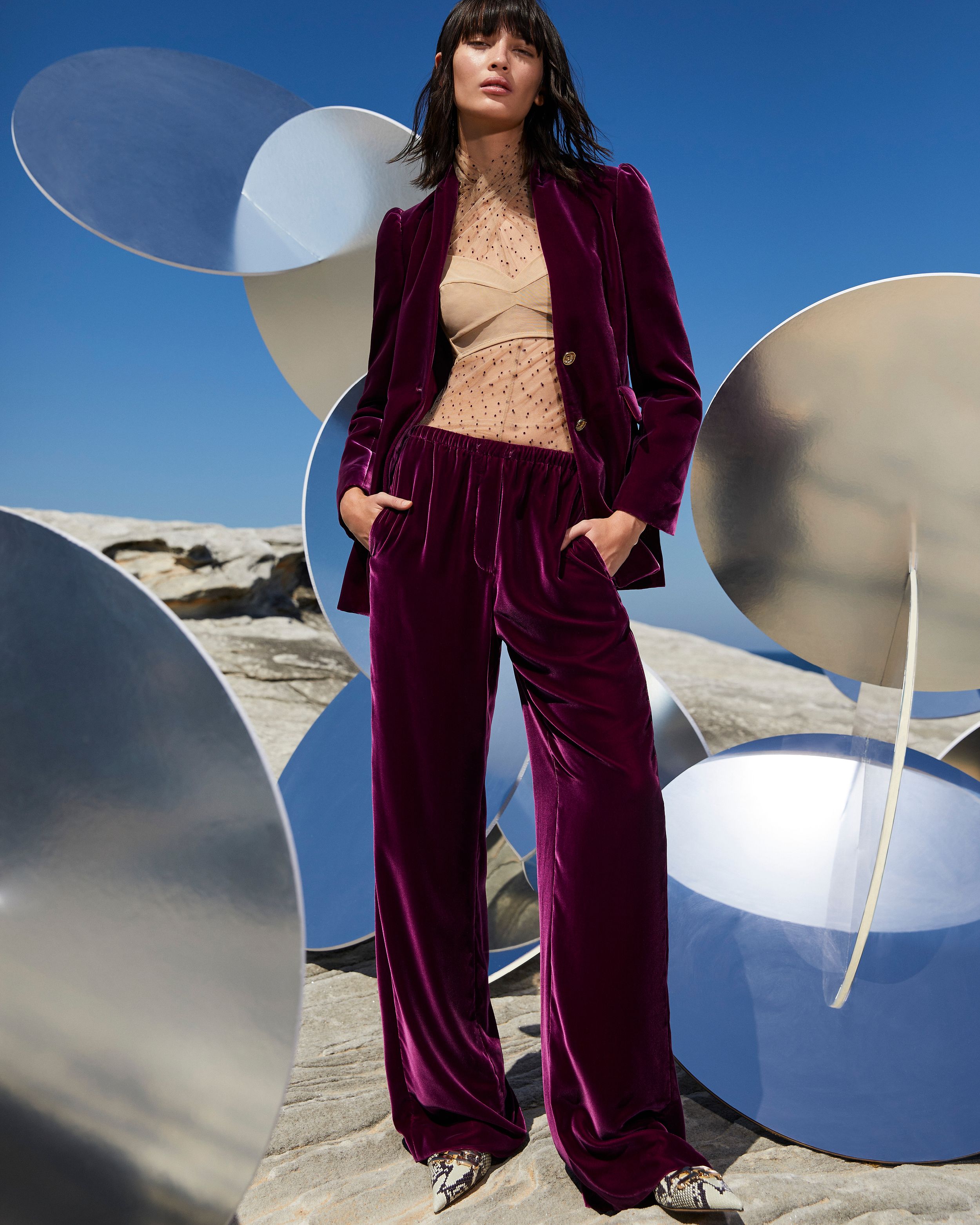 Model standing amongst mirrored sculptures outdoors wearing a mulberry-coloured velvet suit