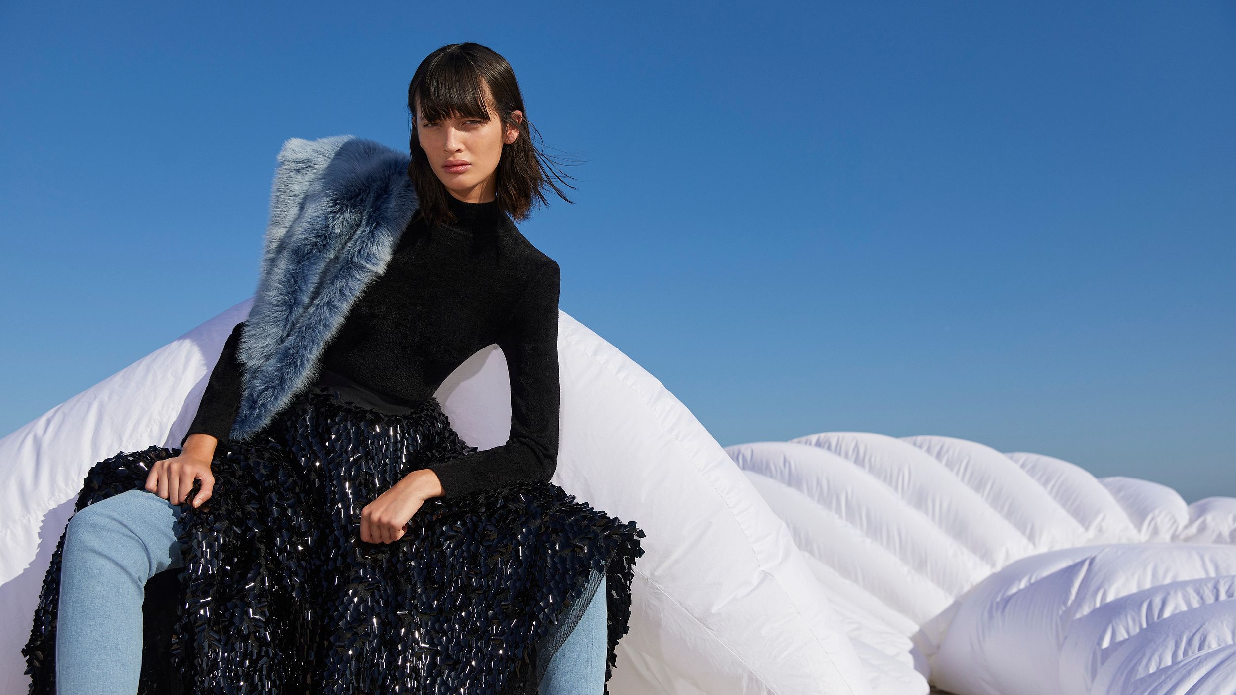 Model sitting on white inflatable structure wearing a black top and sequined skirt with blue boots and Toscana scarf