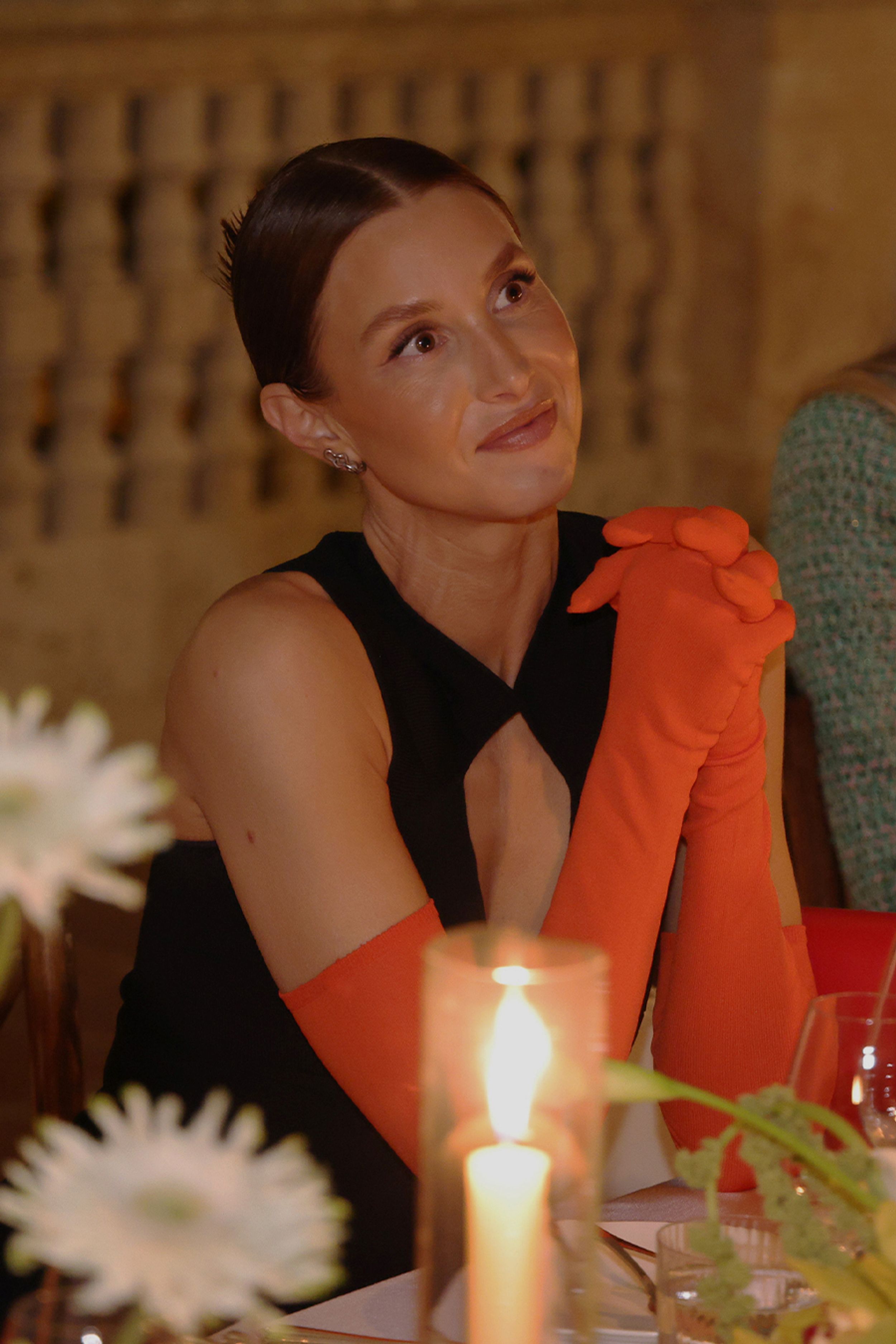 Whitney Port sitting at the table wearing a black sleeveless dress and orange opera gloves