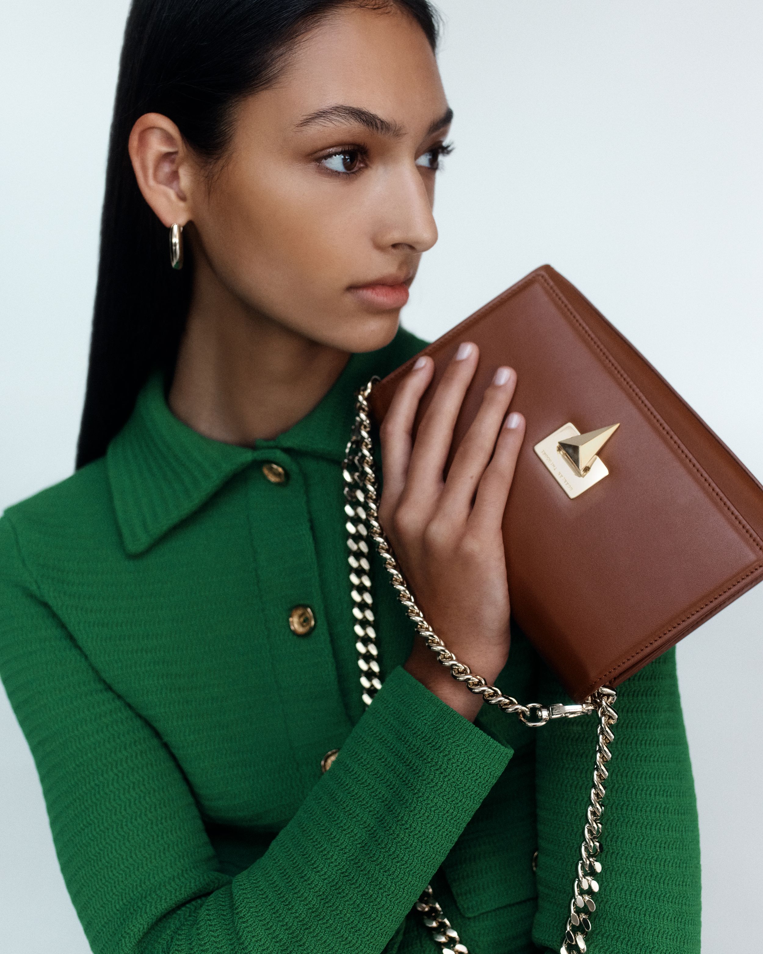 Model looking to the side holding up a brown leather bag with gold hardware, wearing a green collared jacket