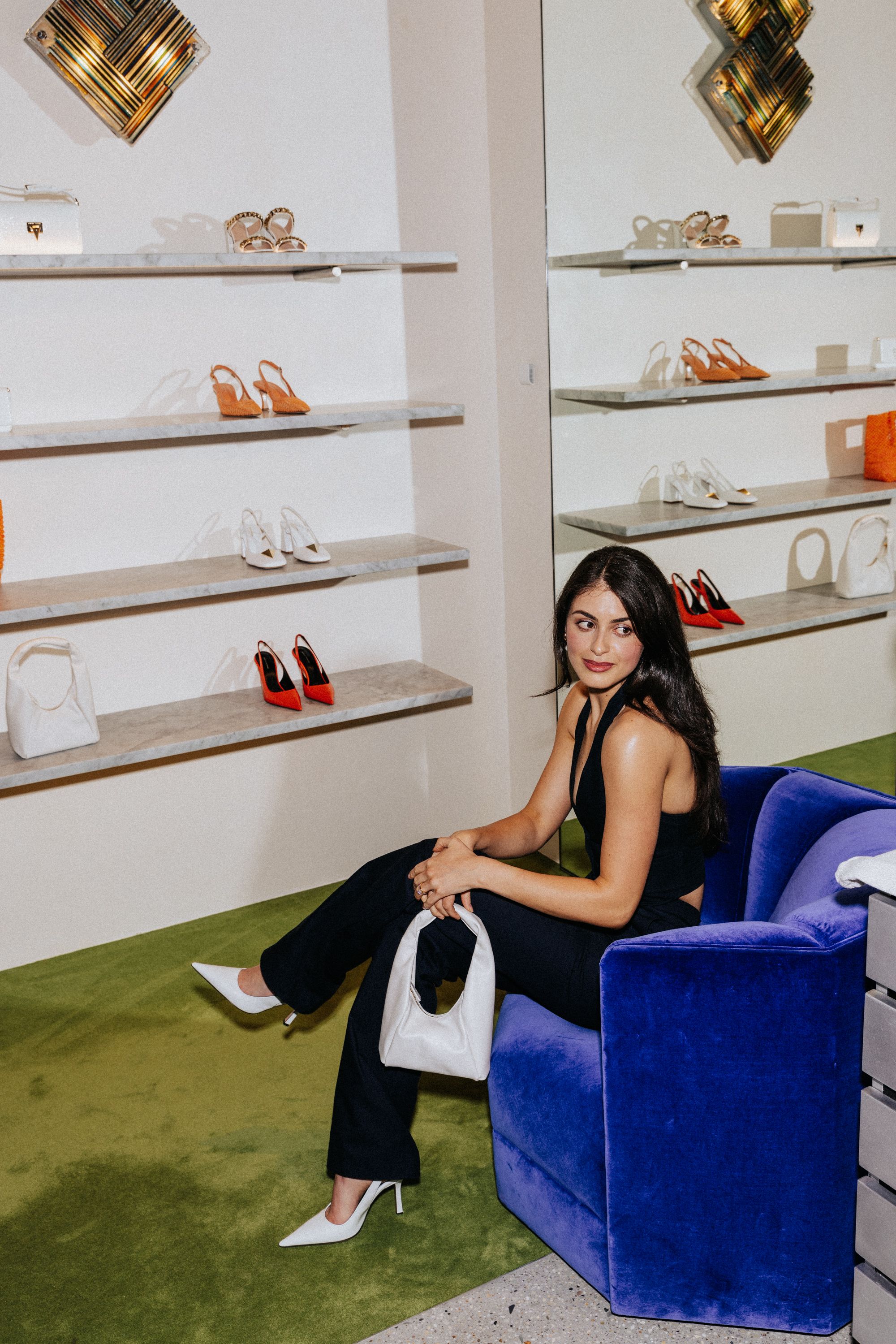 Woman sitting in blue velvet lounge chair next to display wall of shoes and bags