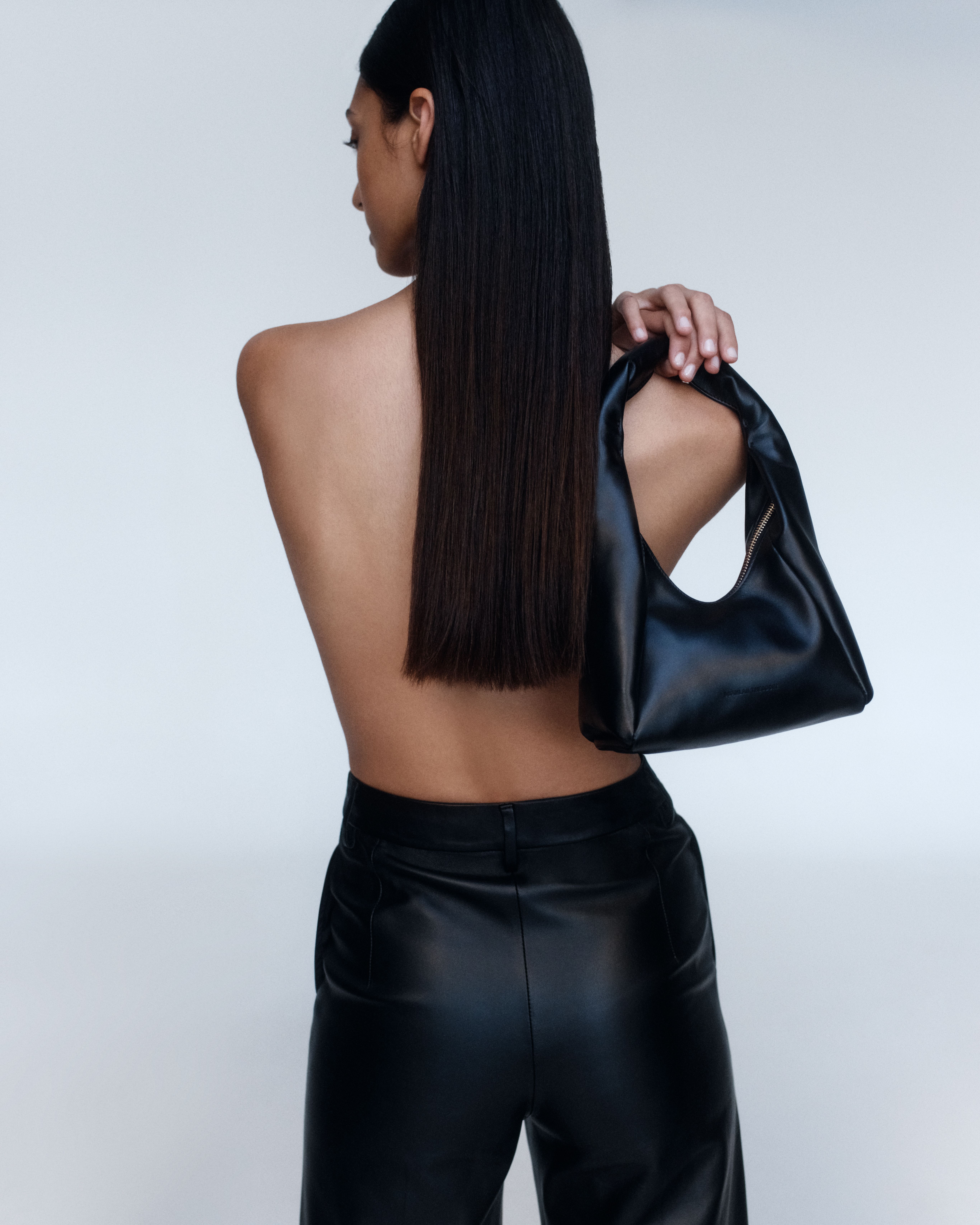 Model facing the wall with long black hair with a small black leather bag draped over her right shoulder