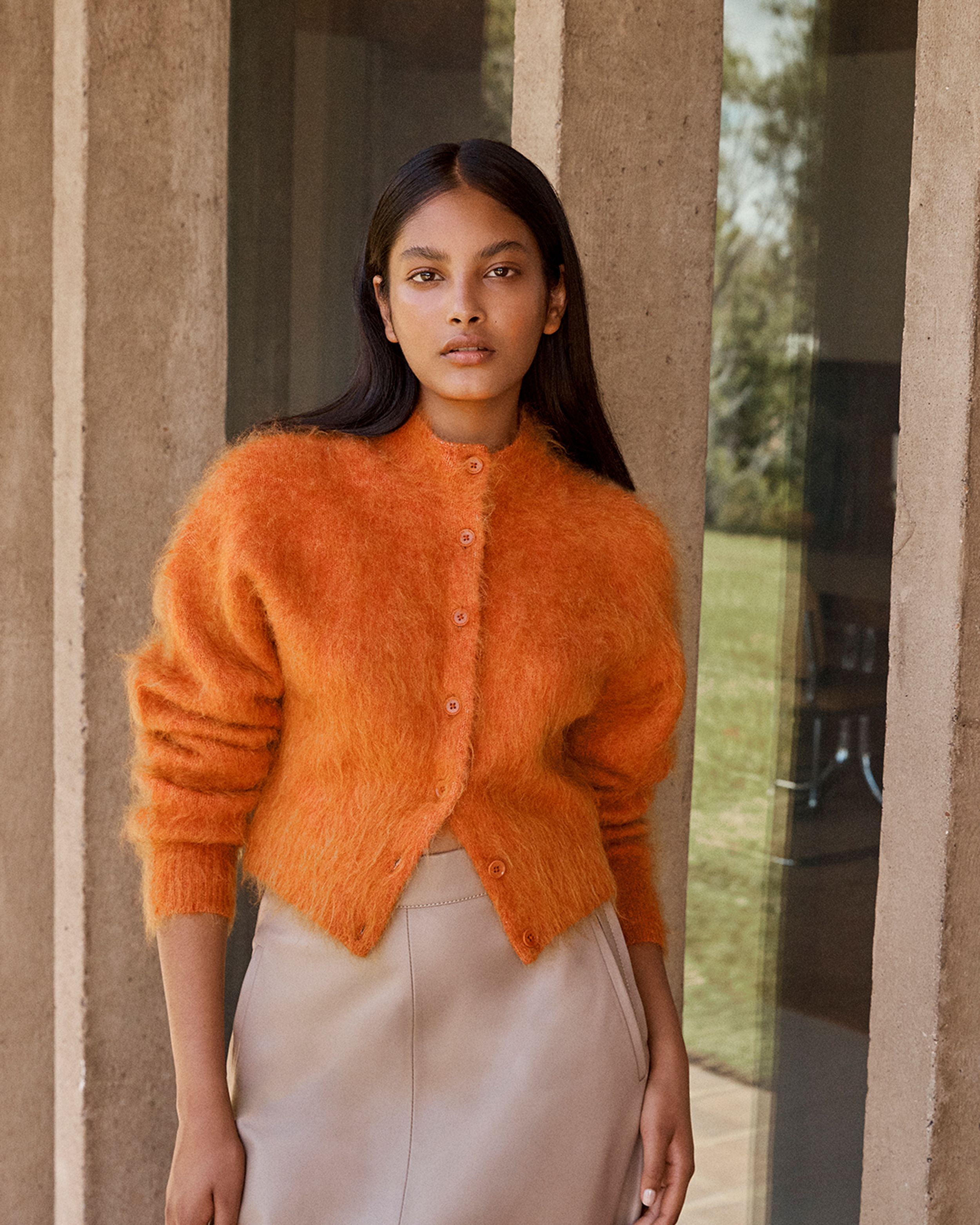 Brunette model standing outside next to a window wearing an orange knit cardigan