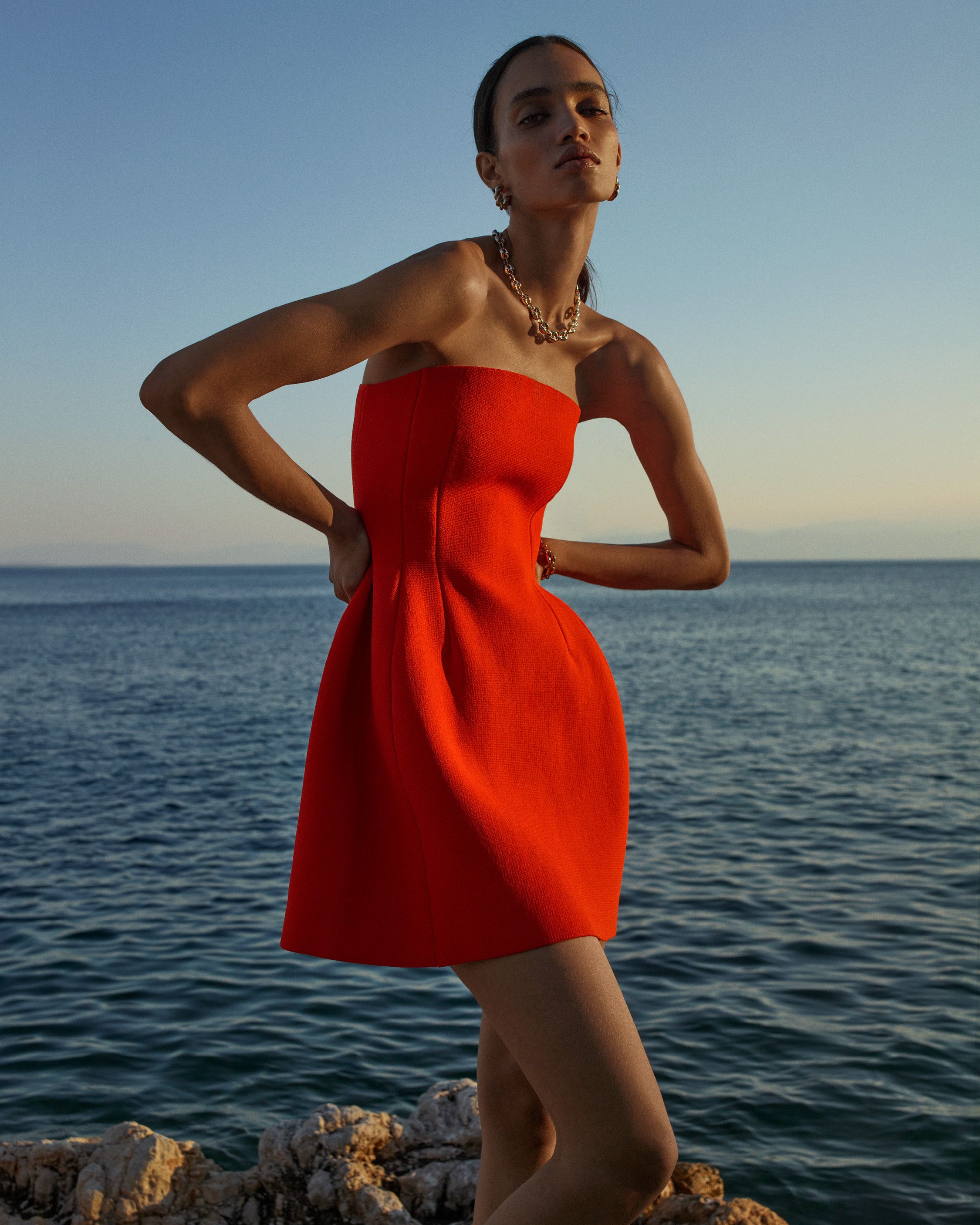 Model standing with hands on hips in front of the ocean wearing a orange strapless mini dress