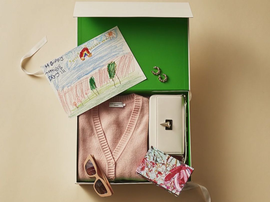 Flatlay of an open green gift box with pink cardigan, sunglasses, white bag, gift card and child's drawing