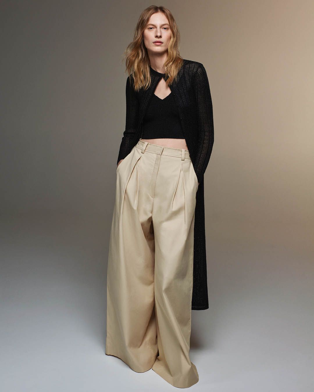 Julia Nobis standing with hands in pockets wearing a matching black singlet and cardigan with beige trousers