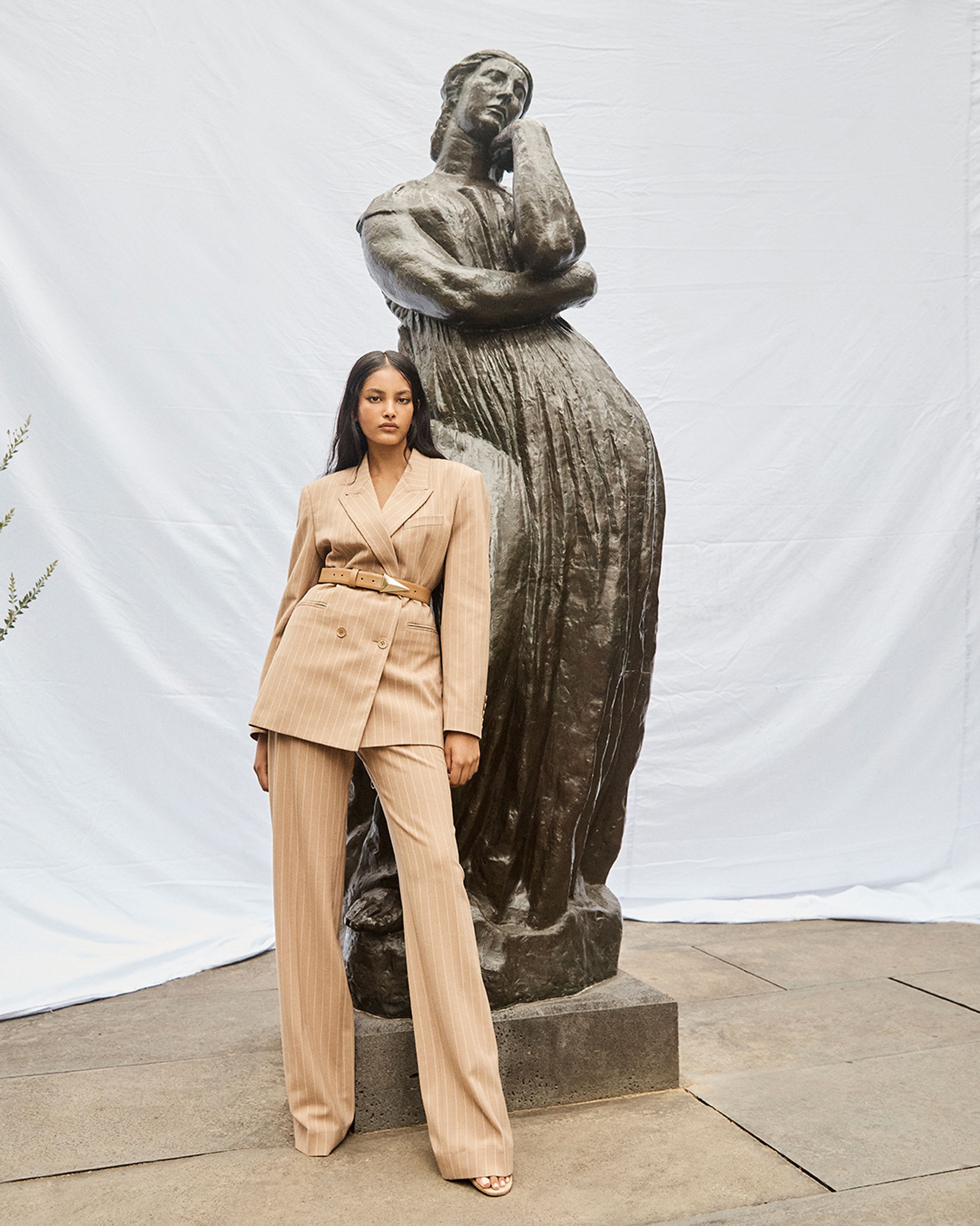 Model wearing a beige pantsuit standing in front of a bronze sculpture