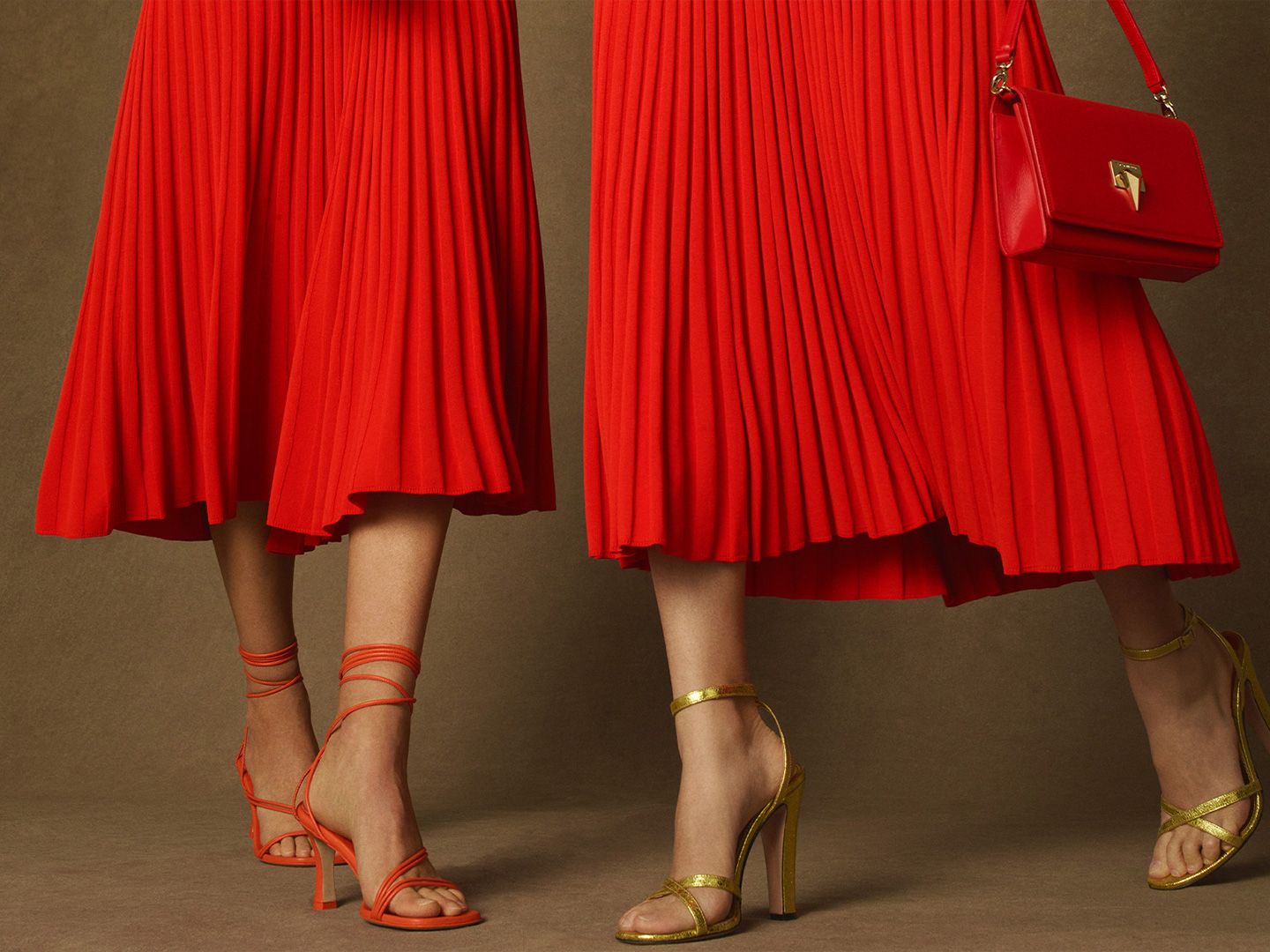 Two models from the legs down both wearing red pleated midi skirts and strappy heels