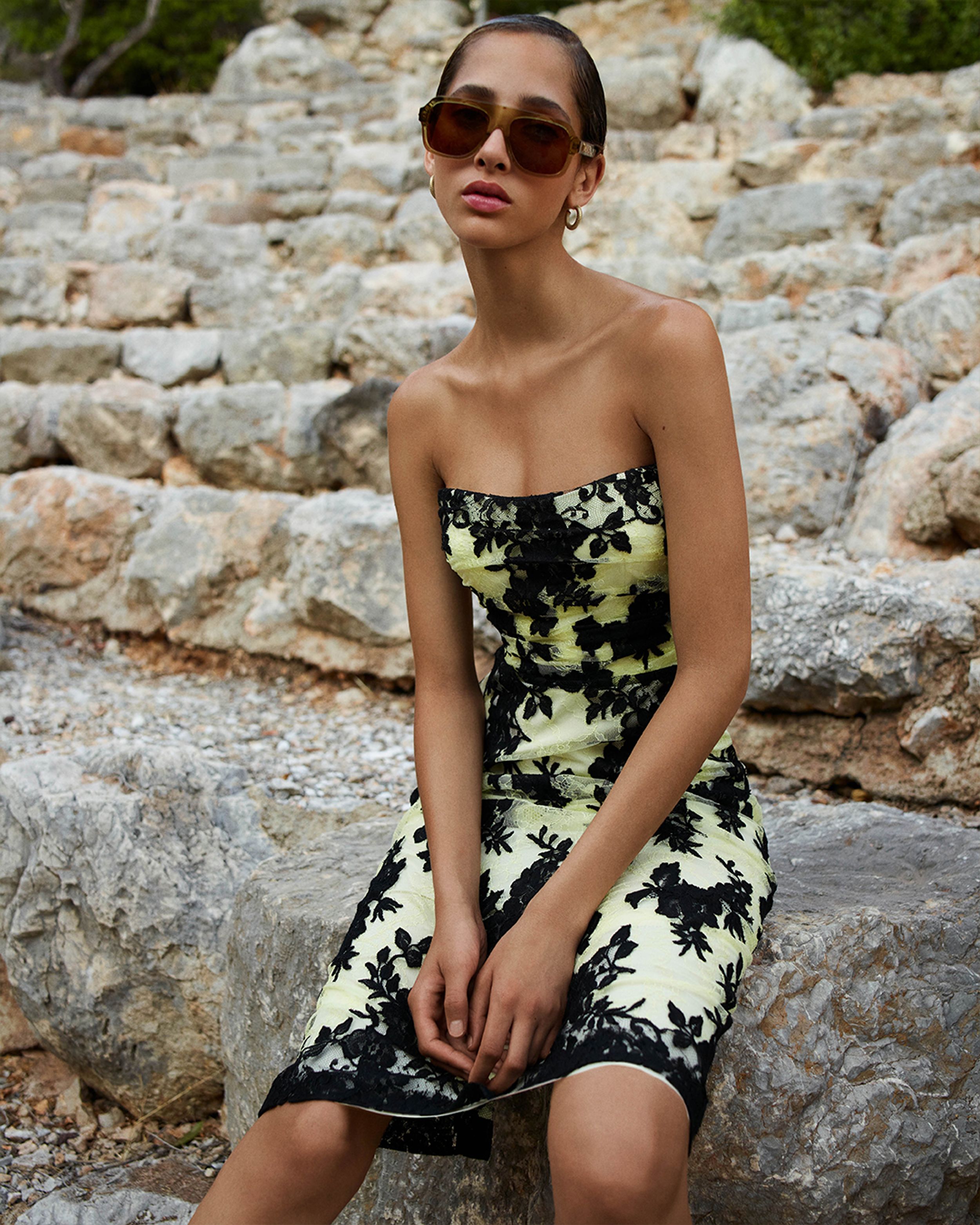 Yasmin Wijnaldum perched on rocks wearing oversized sunglasses and a black and yellow lace mini dress