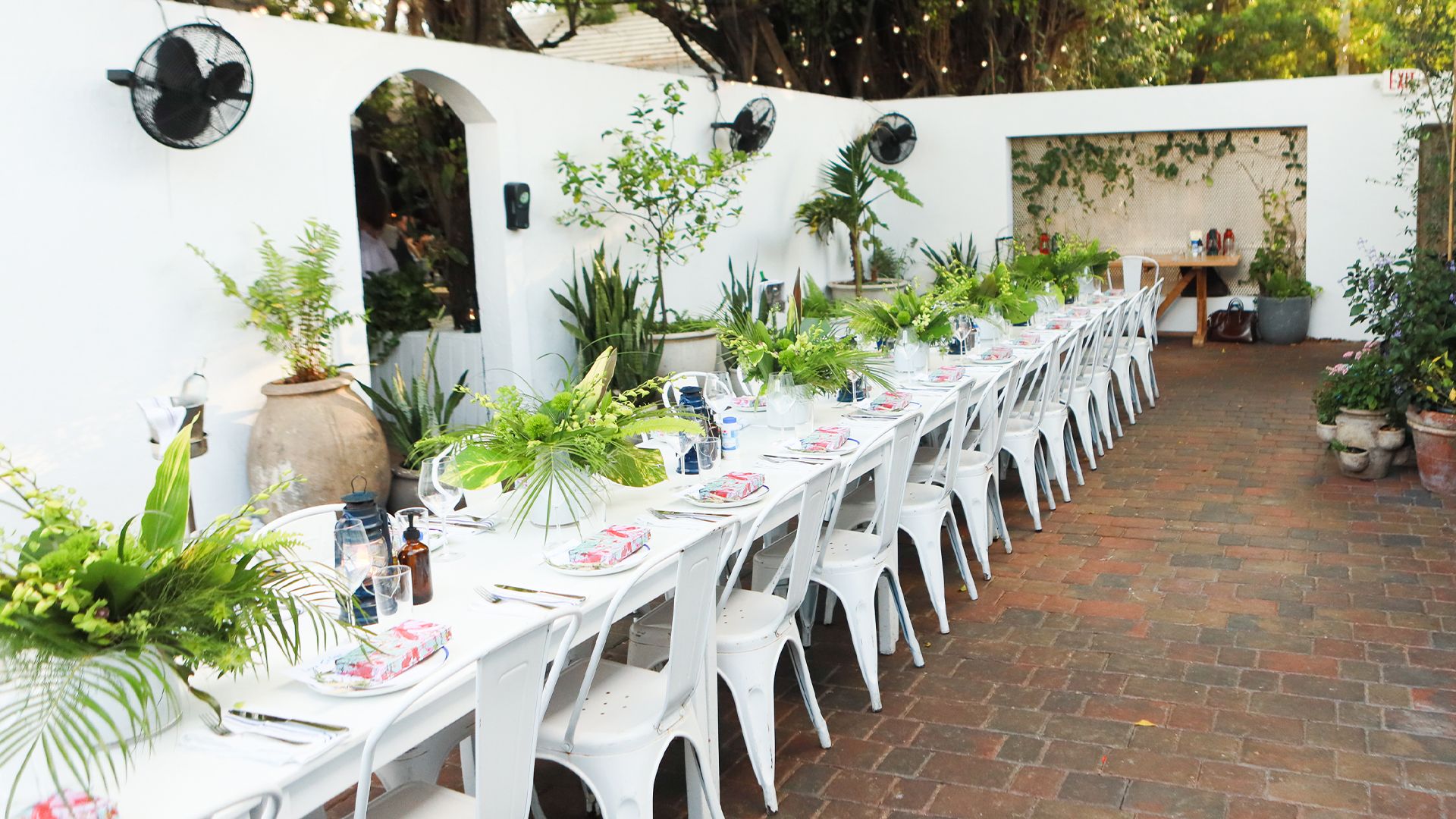 Long white table and chairs in brick courtyard, greenery and plants along table and walls