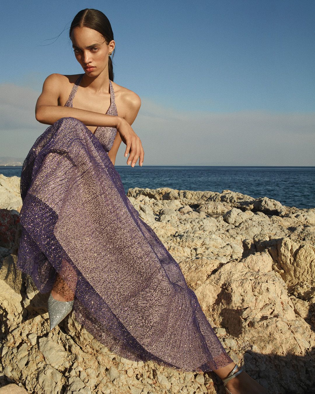 Model wearing a purple sparkly tulle gown sitting on rocks with the ocean in the background