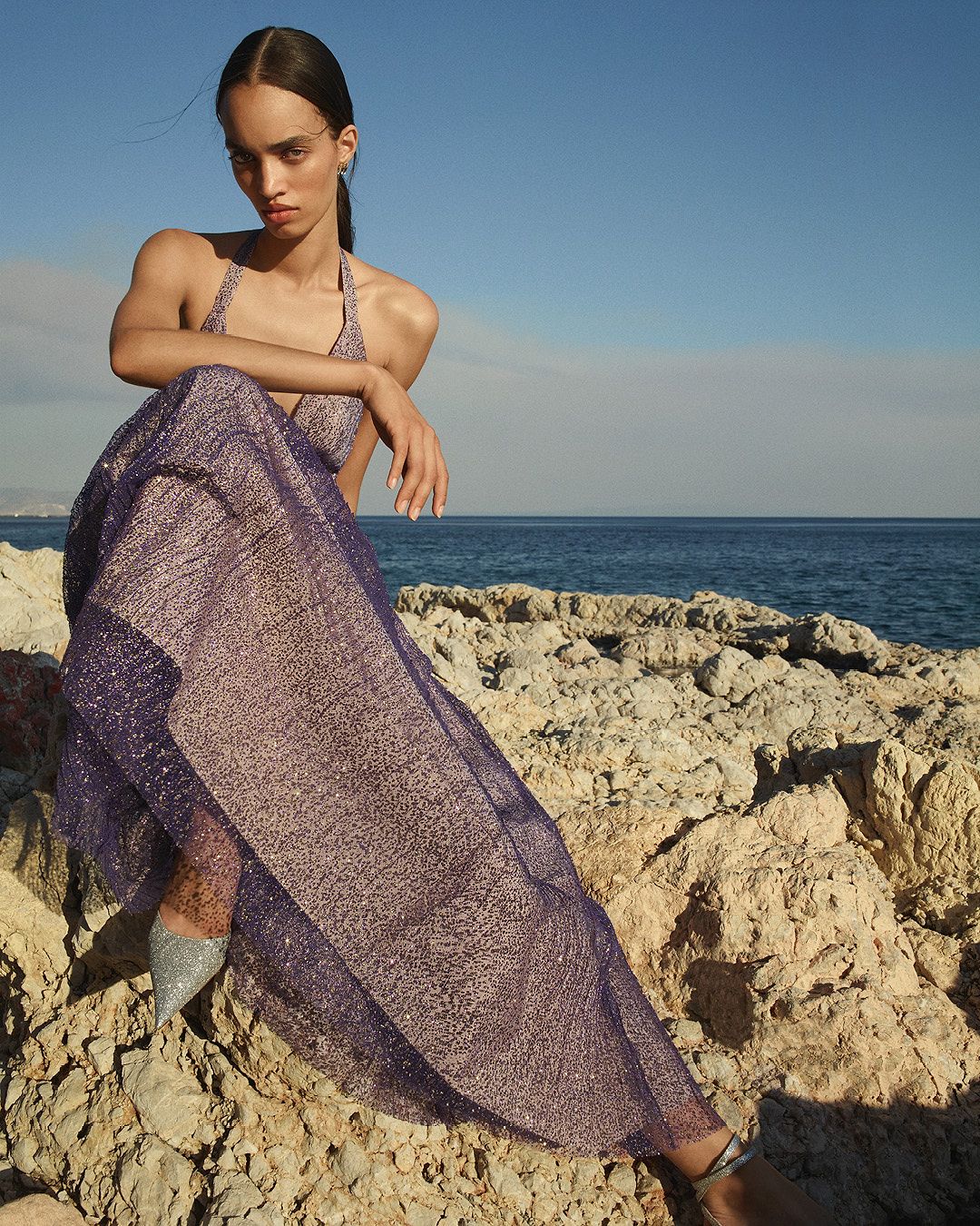 Model wearing a purple sparkly tulle gown sitting on rocks with the ocean in the background
