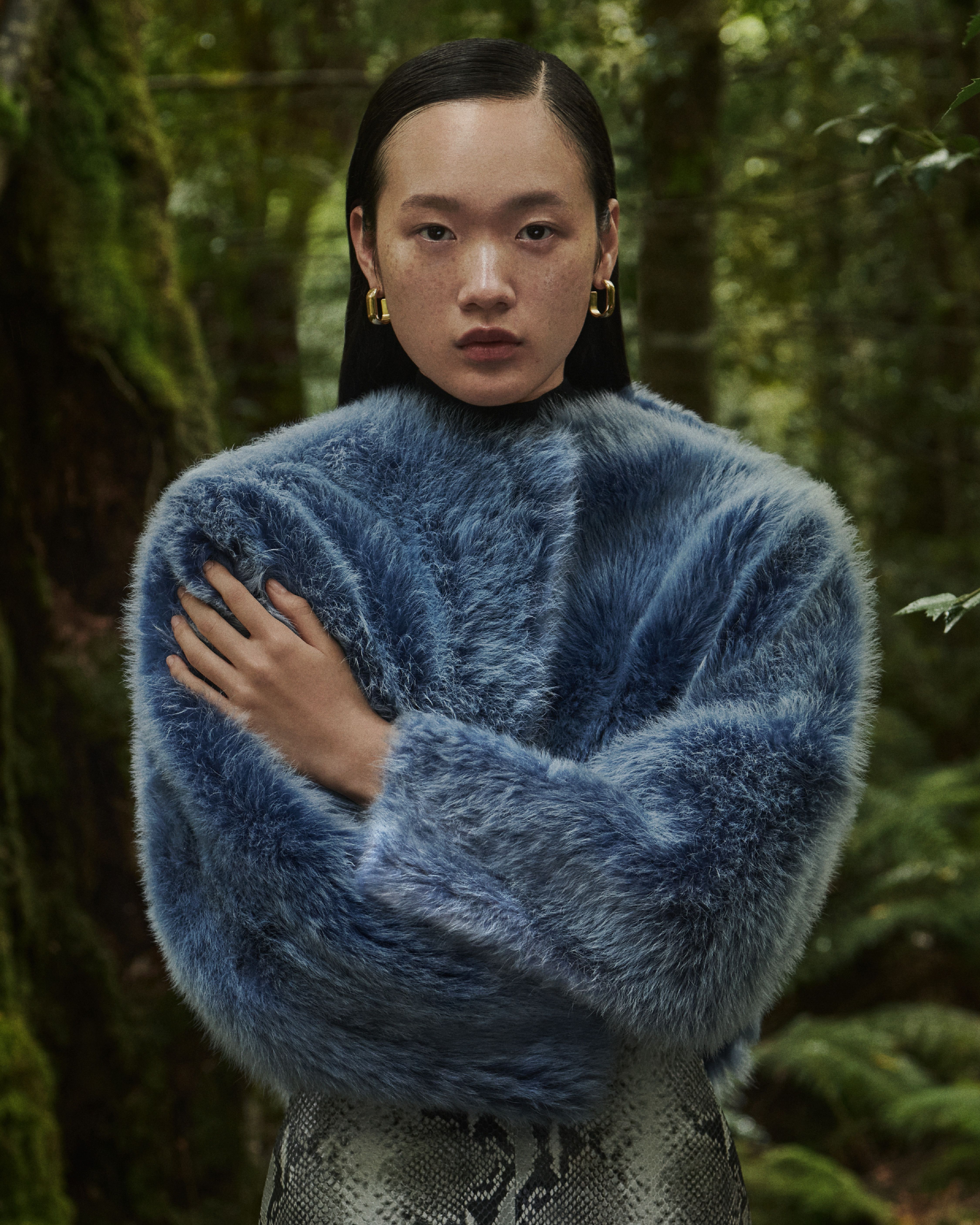 Mid shot of Venus He standing in the forest with arms crossed wearing a blue Toscana shearling jacket