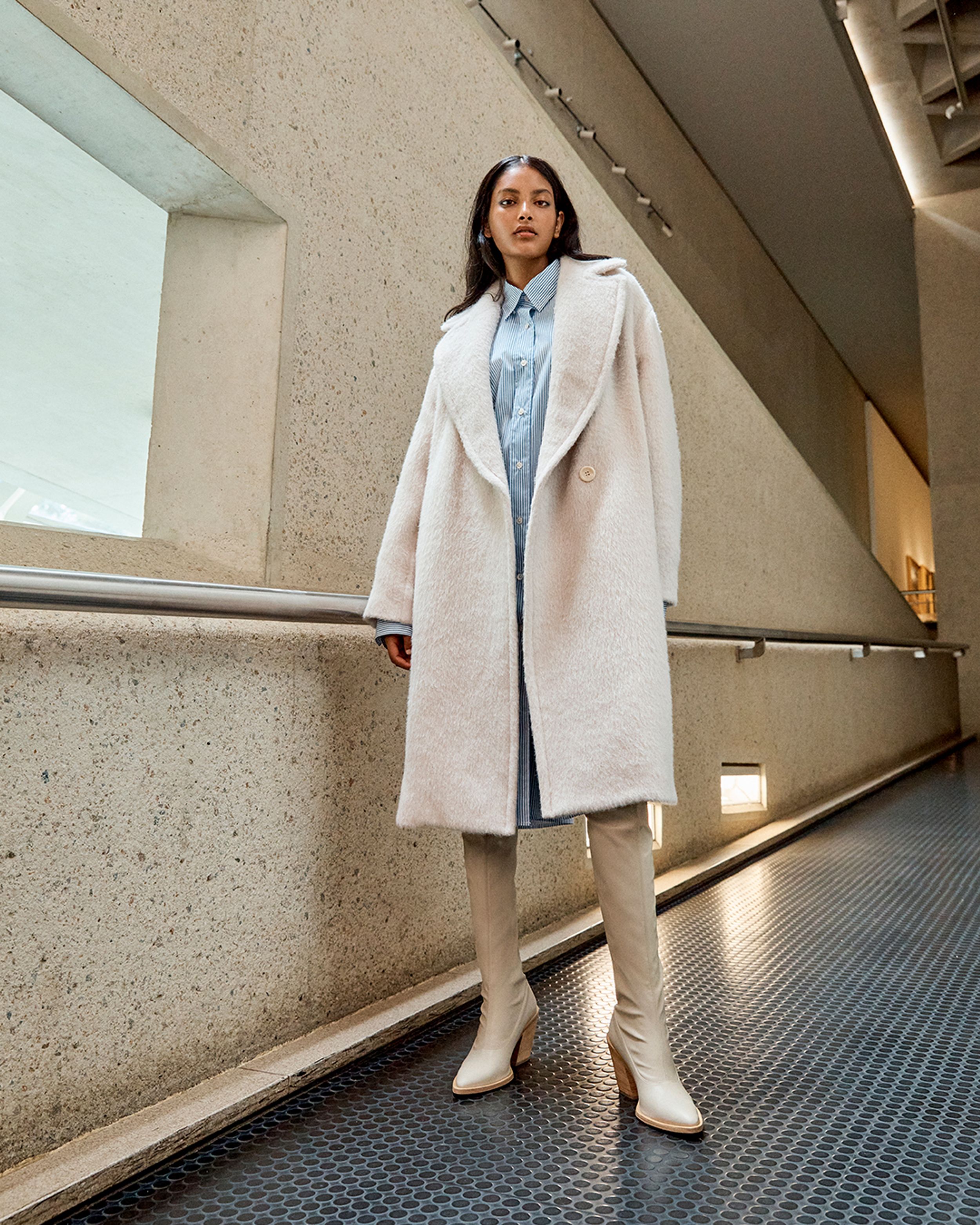 Model standing in a hallway by a window wearing a blue striped shirt dress and cream coat