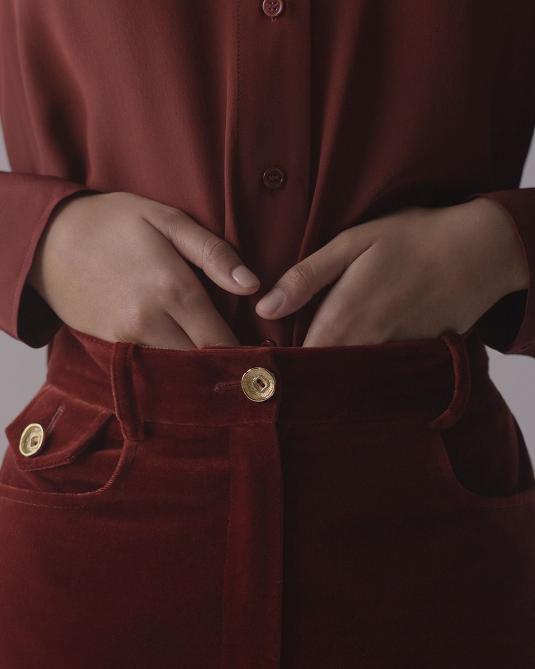 Woman’s hands tucking into red velvet trousers, gold button on front of pants and pocket. 