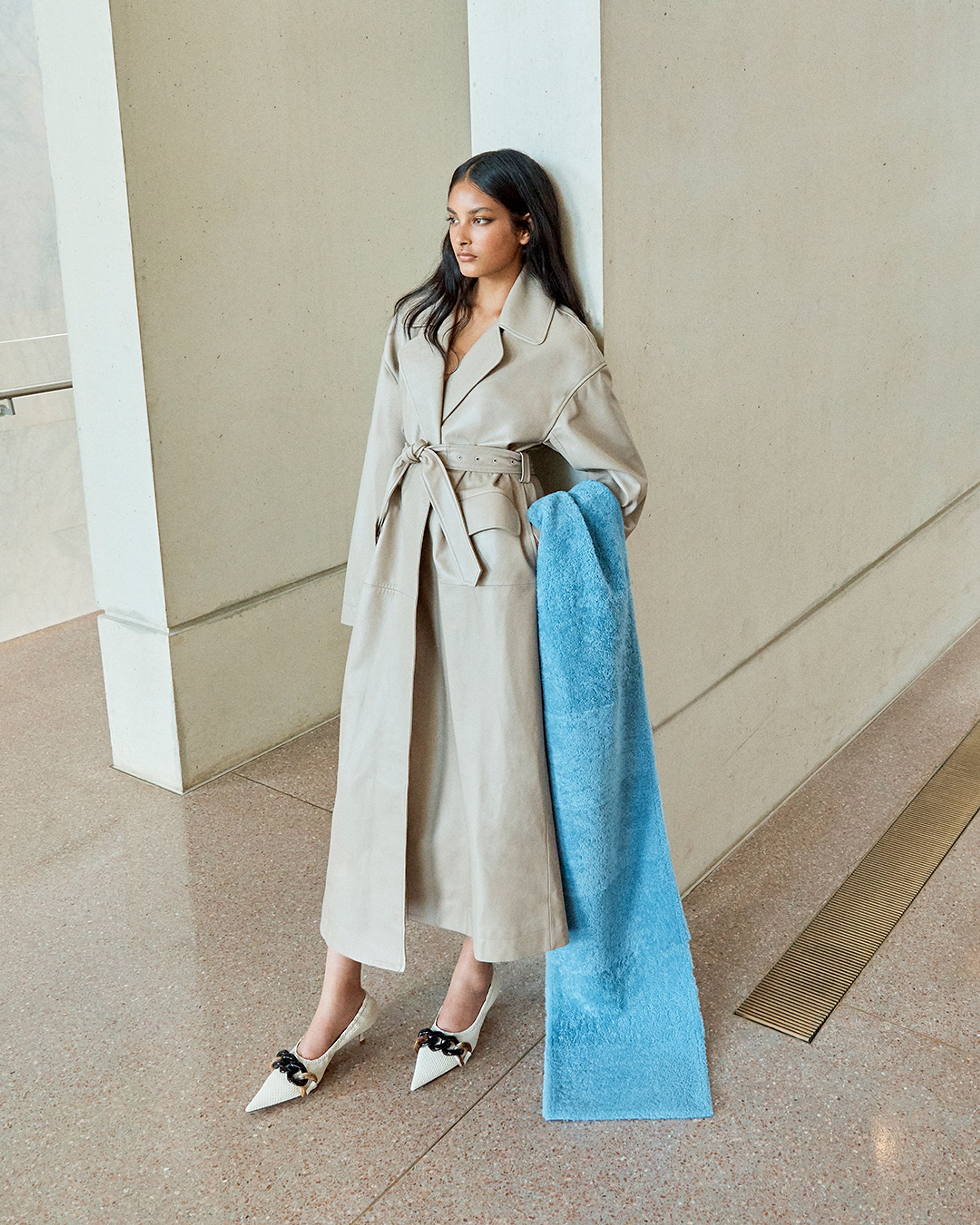 Brunette model leaning against the wall wearing a neutral leather trench and blue scarf