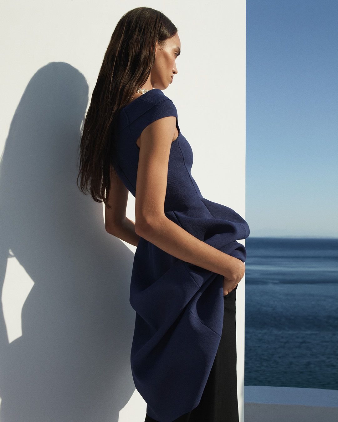 Model facing the side with hands in pockets wearing a dark blue cold shoulder dress