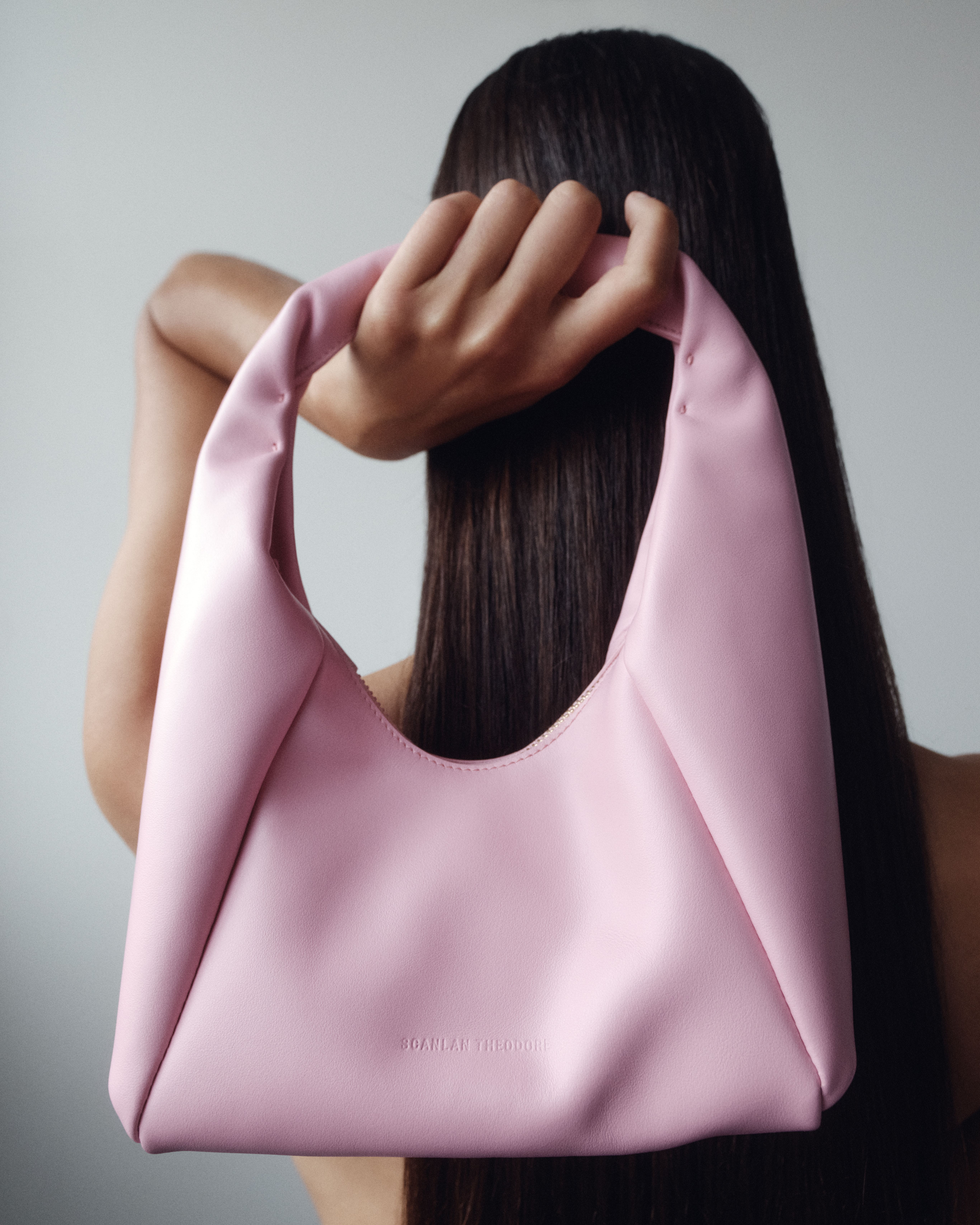 Model facing the wall with long straight brown hair holding a light pink leather bag behind her head