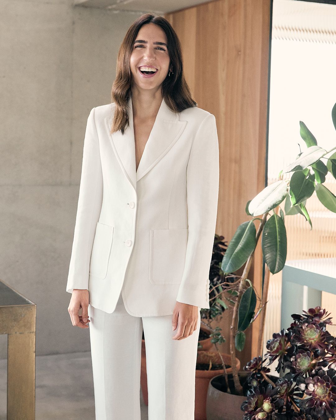 Bianca Marchi smiling in a white pantsuit posing next to a potted plant