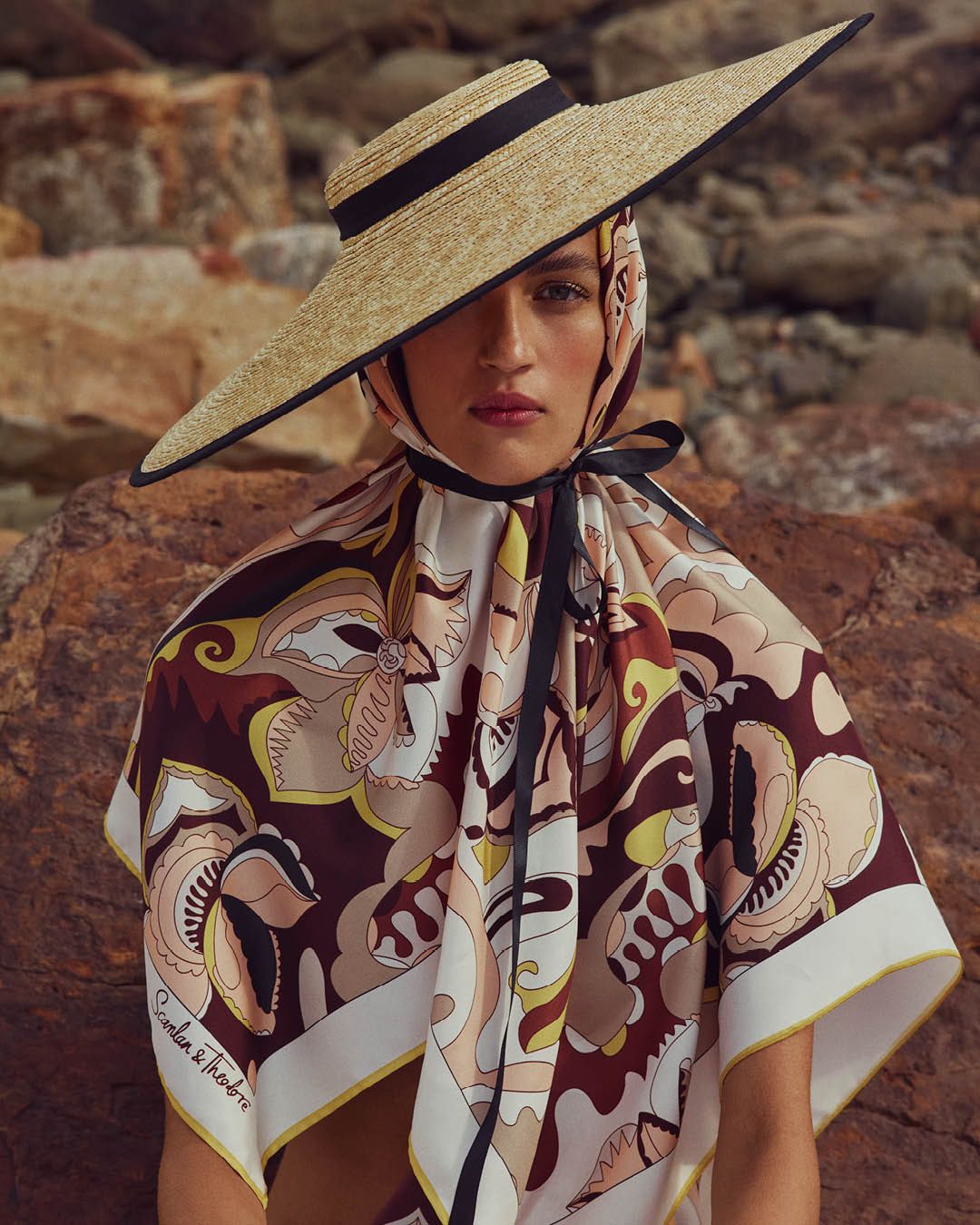 Model gazing at the camera wearing straw hat with a brown, yellow and white scarf over her head and shoulders