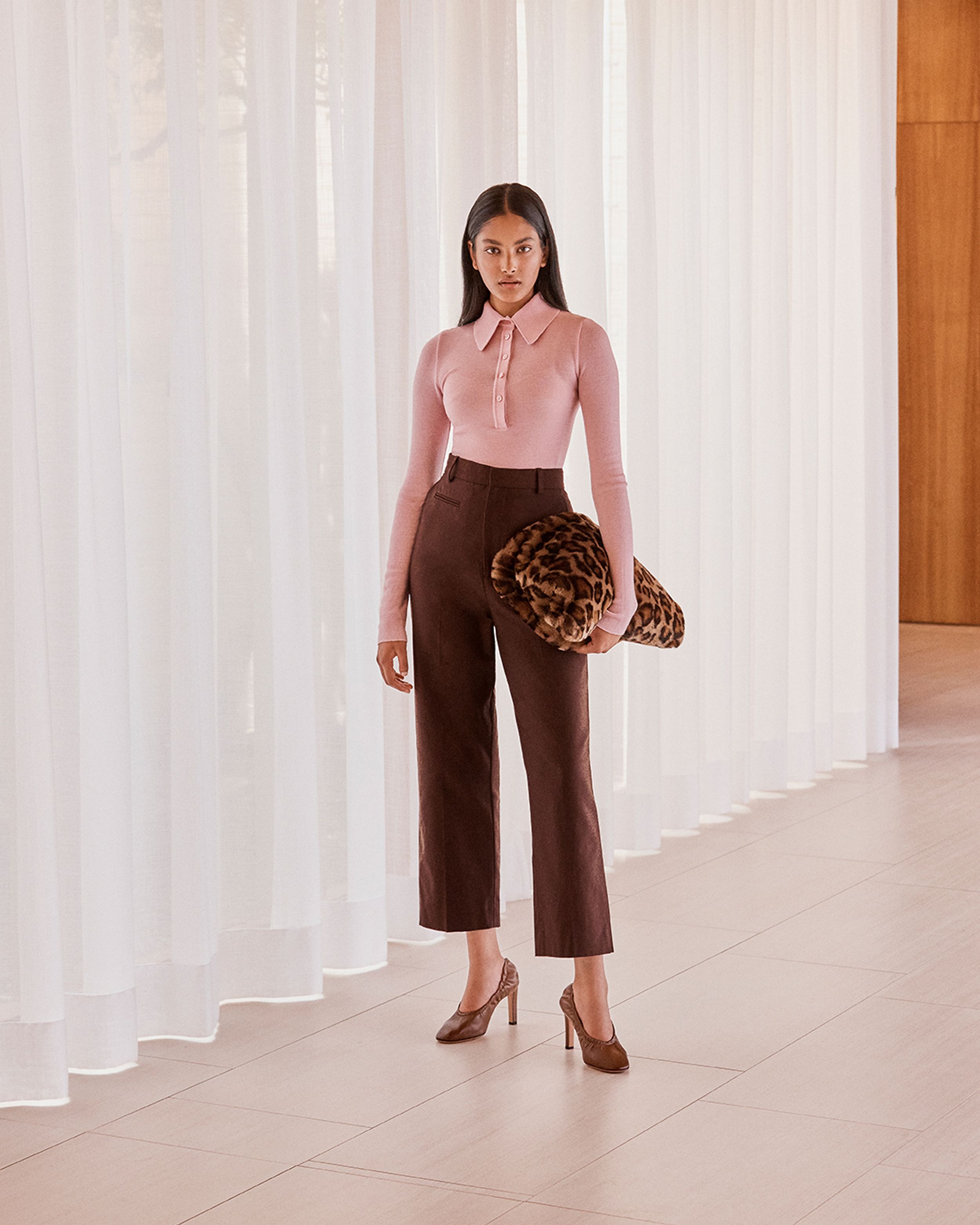 Brunette model standing by a window wearing a pink knit shirt and burgundy trousers