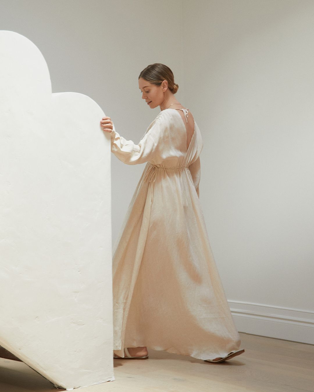 Hayley Bonham with hair in a sleek bun walking away up the stairs wearing a long maxi dress in neutral colourway