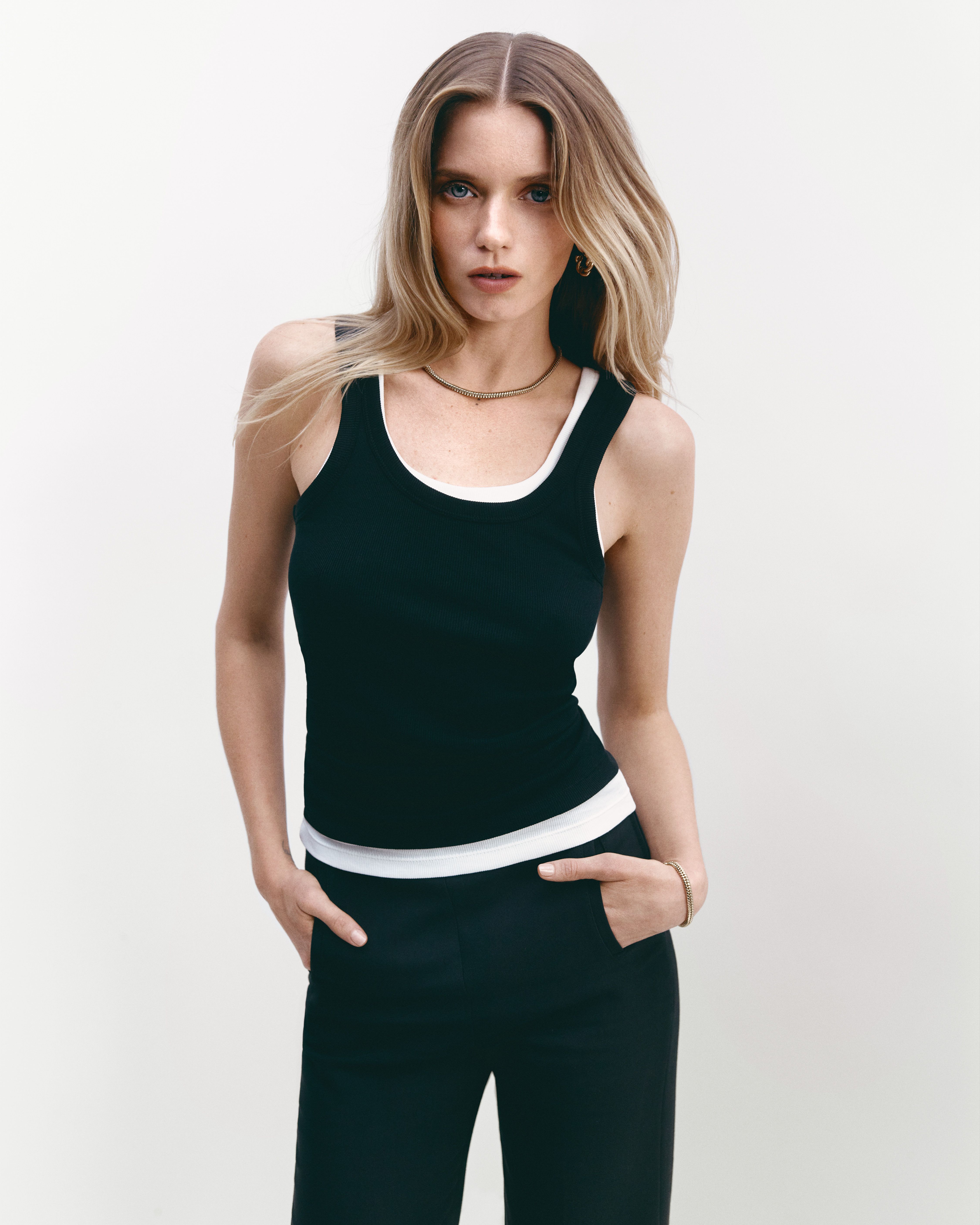 Abbey Lee standing with hands in pockets wearing black and white singlets layered and black trousers