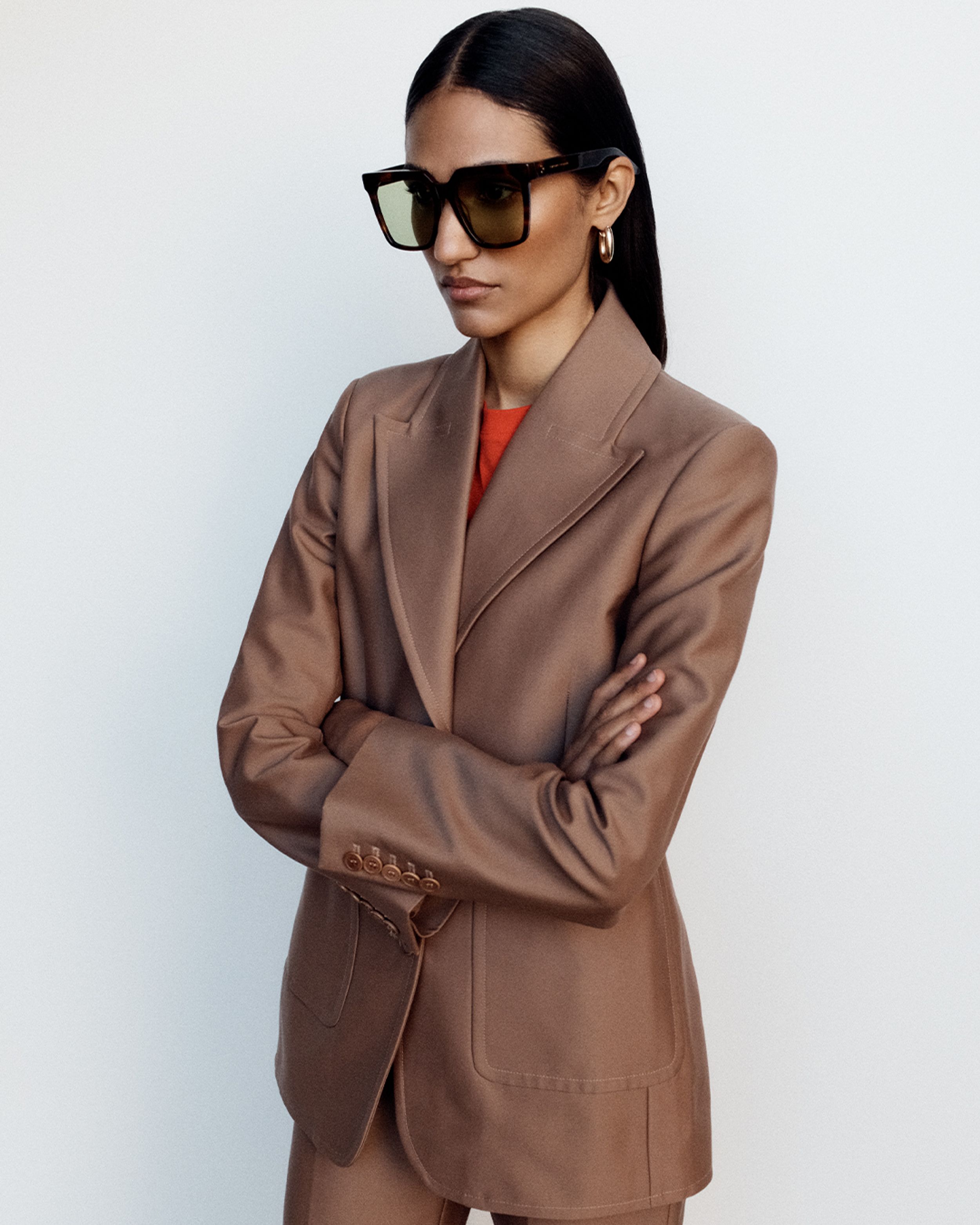 Model posing with arms crossed wearing dark brown oversized sunglasses with a brown pantsuit