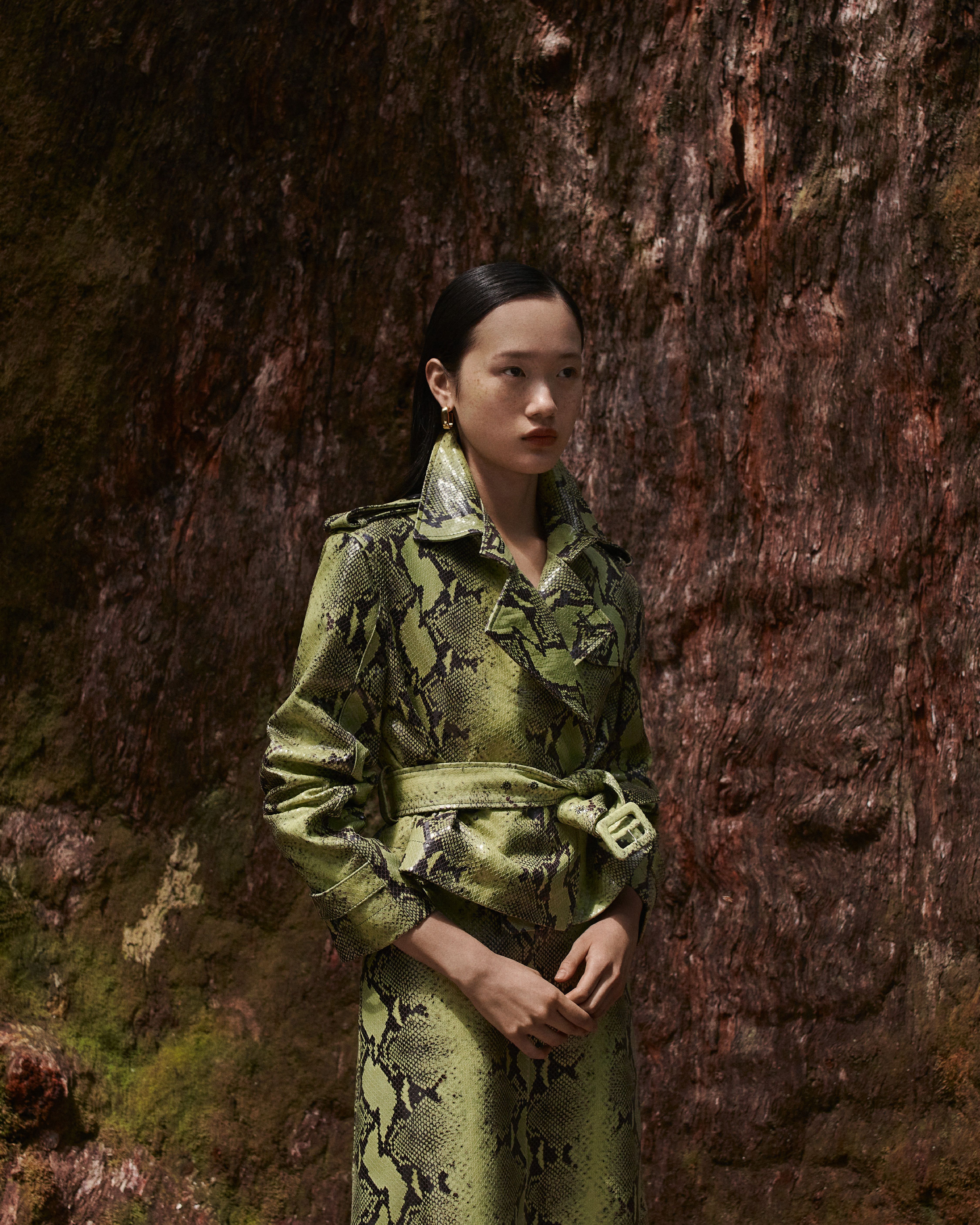 Venus He standing in front of a tree wearing a reptile-print leather trench coat