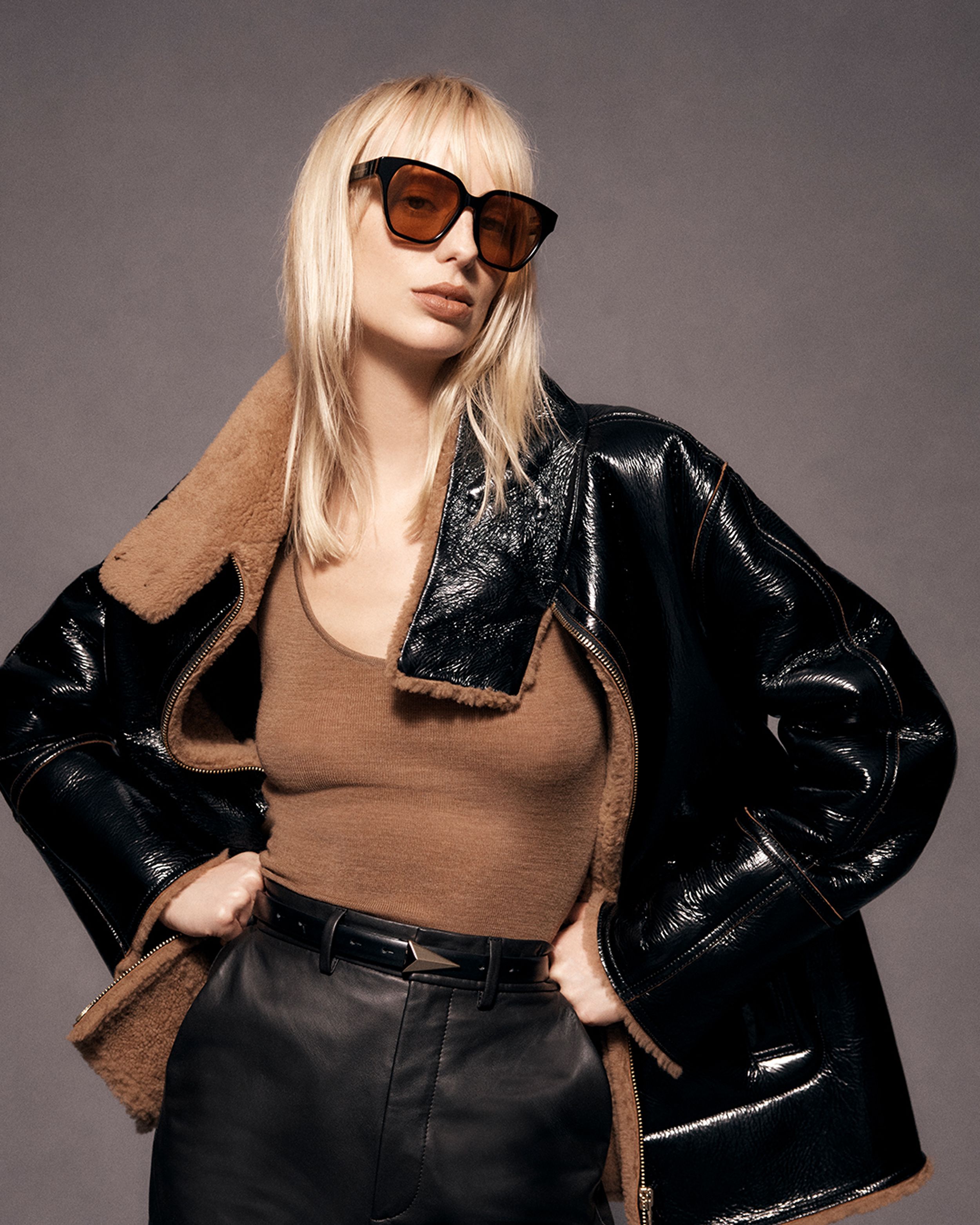 Blonde model posing with hands on hips wearing a black shearling jacket and brown tank top