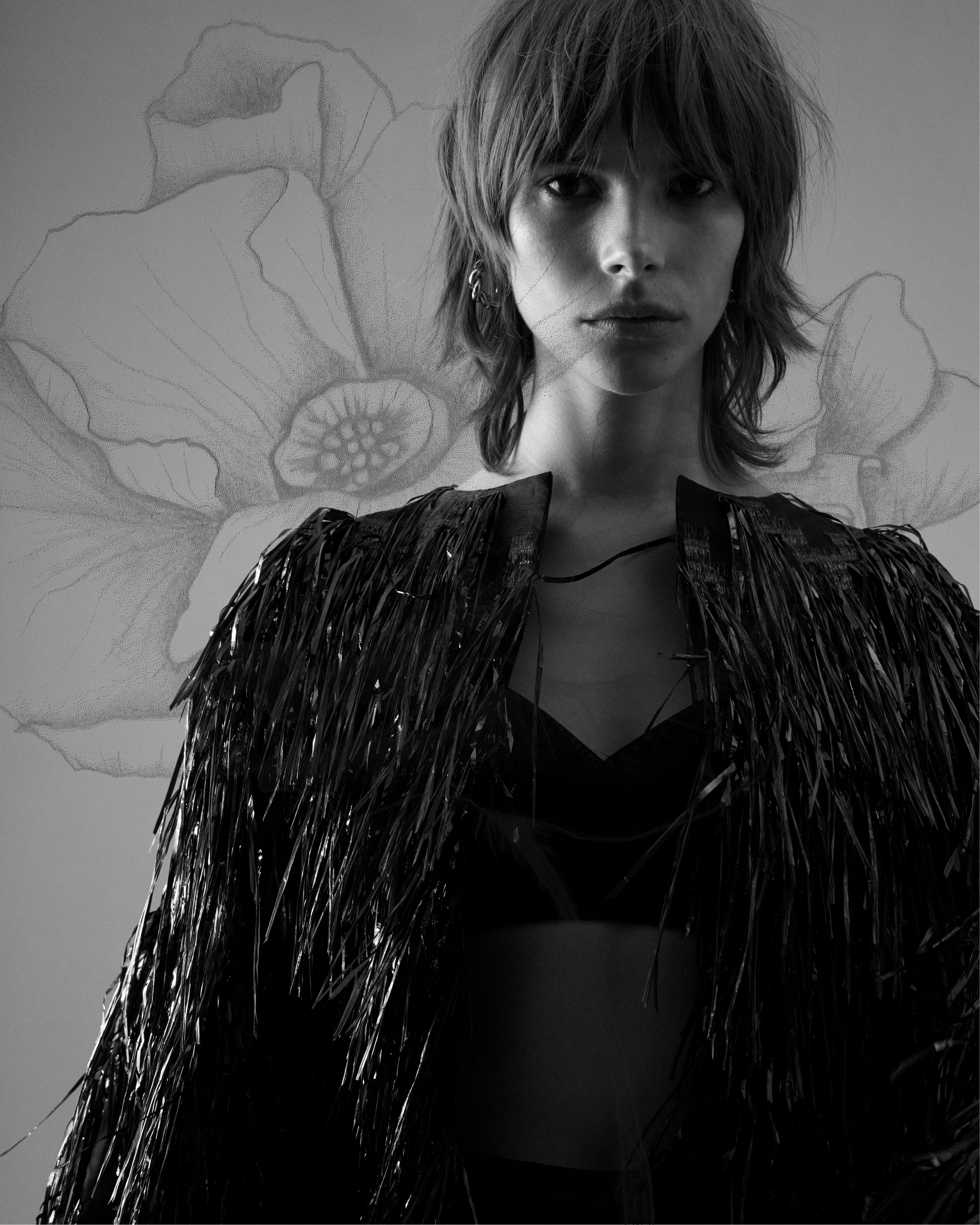 Black and white image of woman wearing a black tinsel fringed jacket and bralette with poppy illustration in background