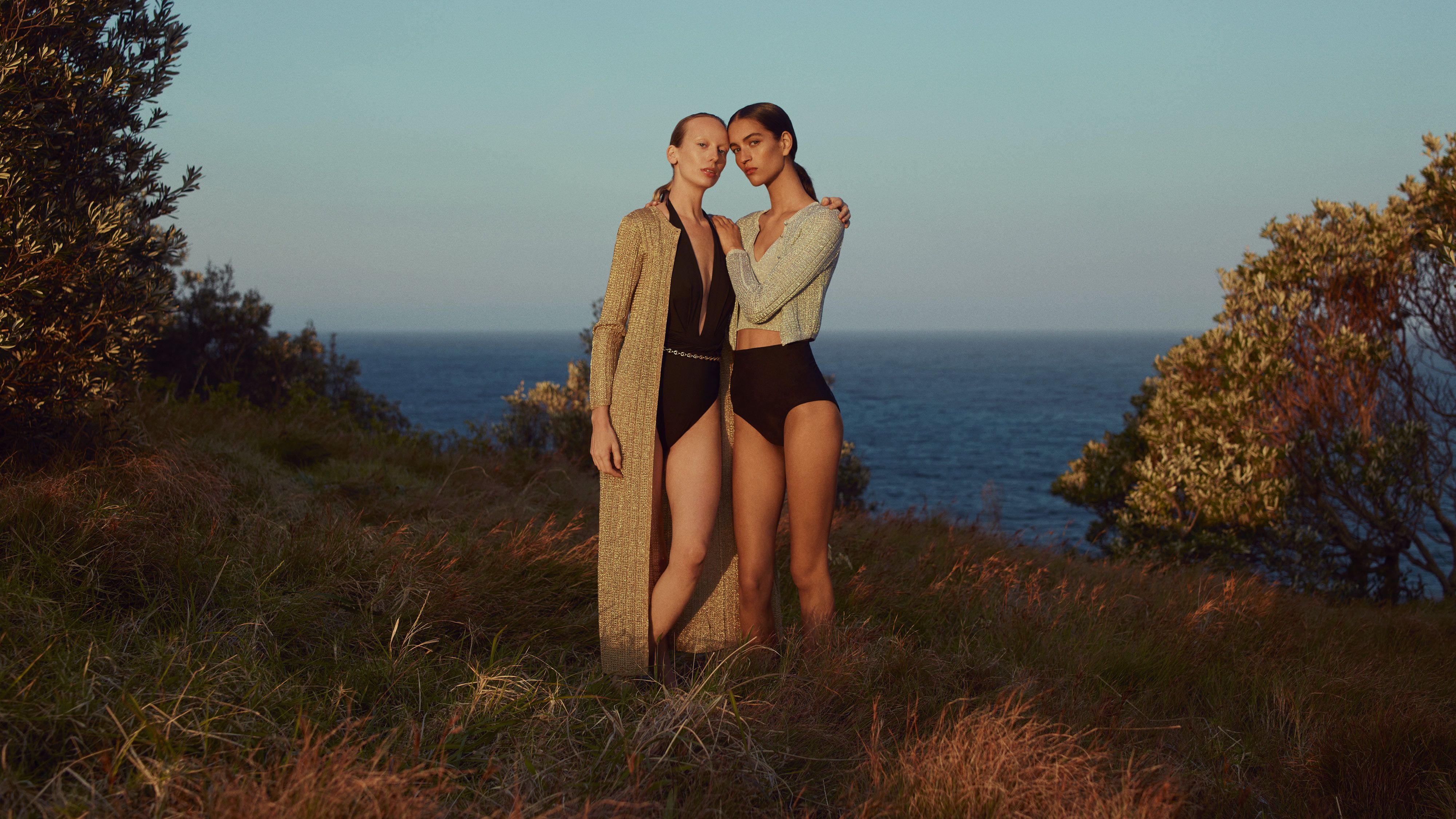 Two models standing on grass with the ocean in the background embracing wearing metallic knit cardigans and black swimwear