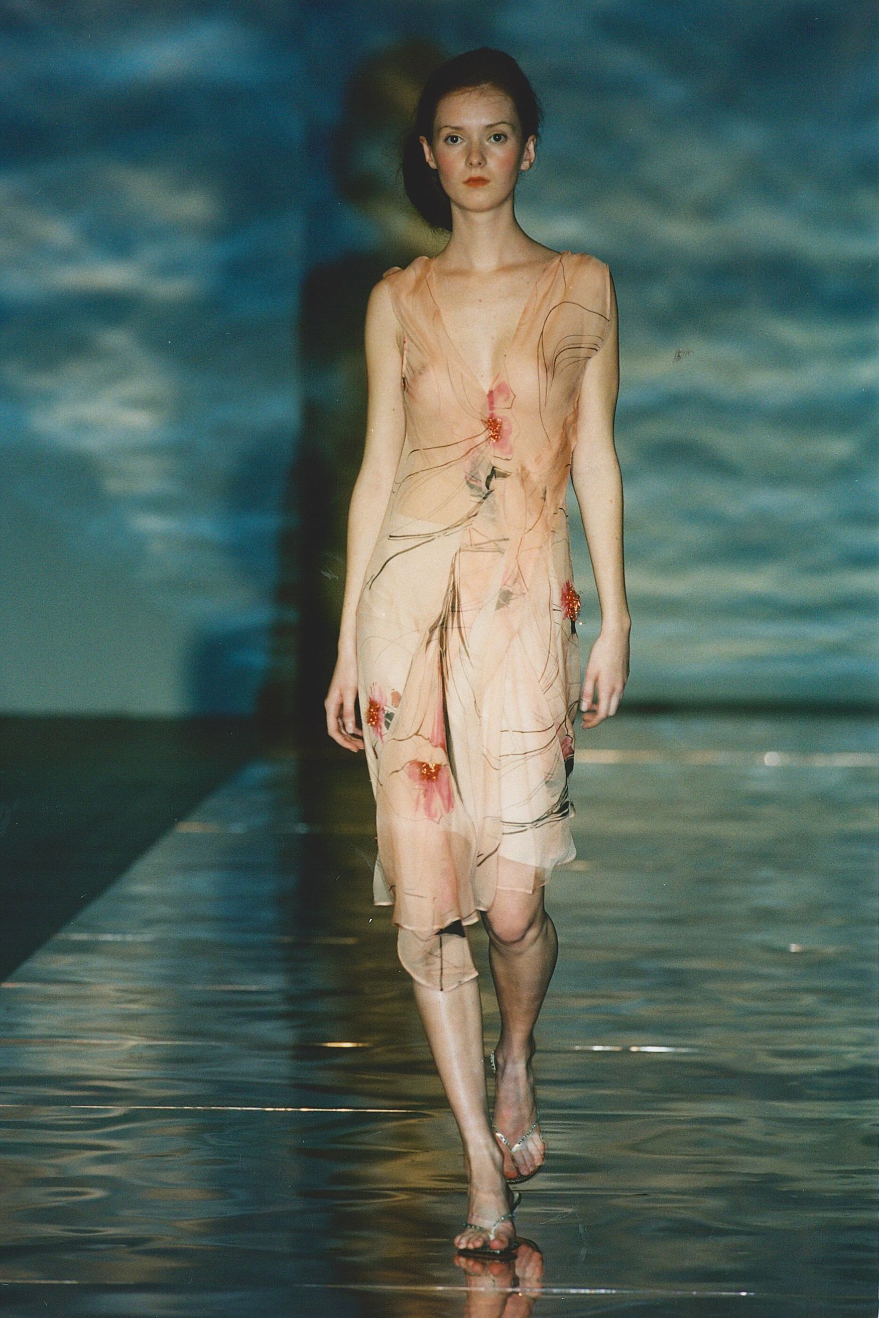 Model walking down the runway in a beige silk draped dress with floral embroidery