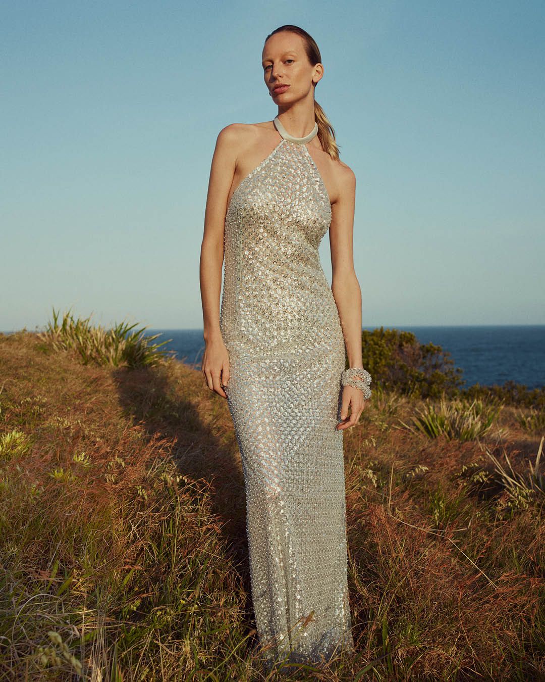Blonde model wearing a silver sequined halterneck evening gown standing on the grass with the ocean in the background