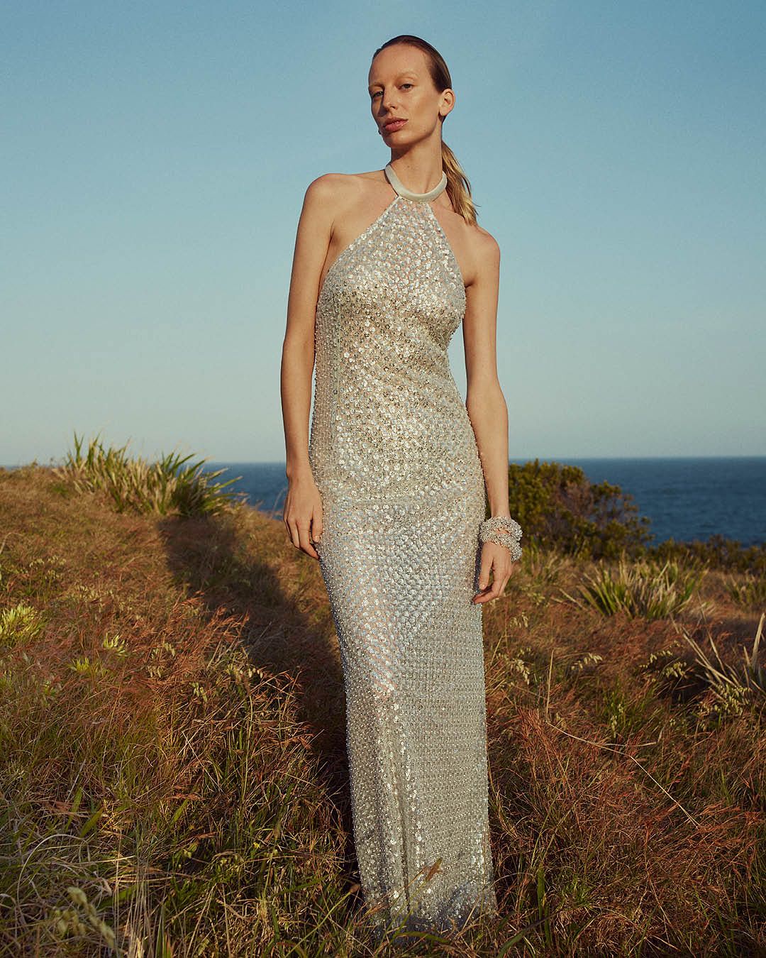Blonde model wearing a silver sequined halterneck evening gown standing on the grass with the ocean in the background