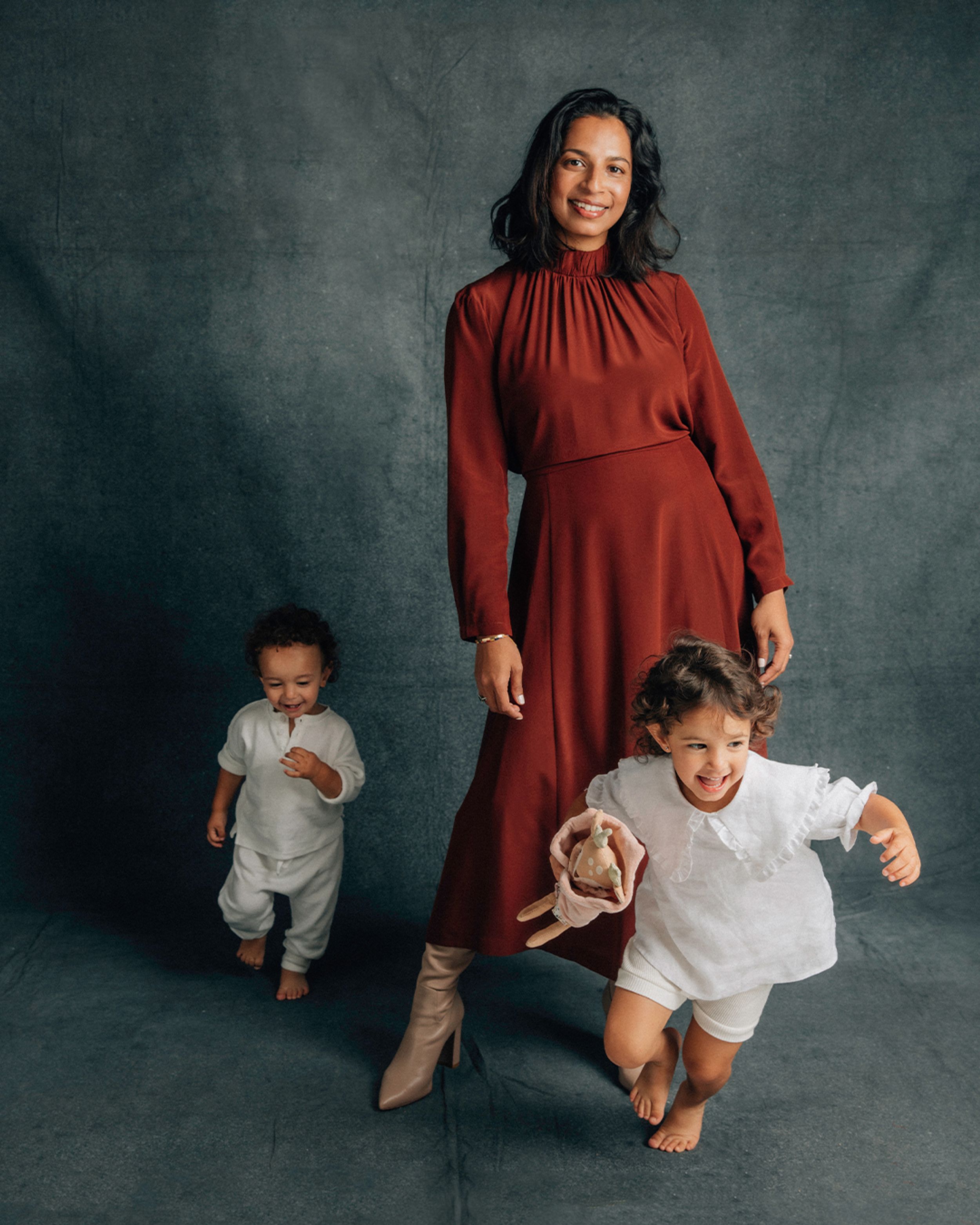 Shamini Rajarethnam smiling wearing a rust silk midi dress while her children run