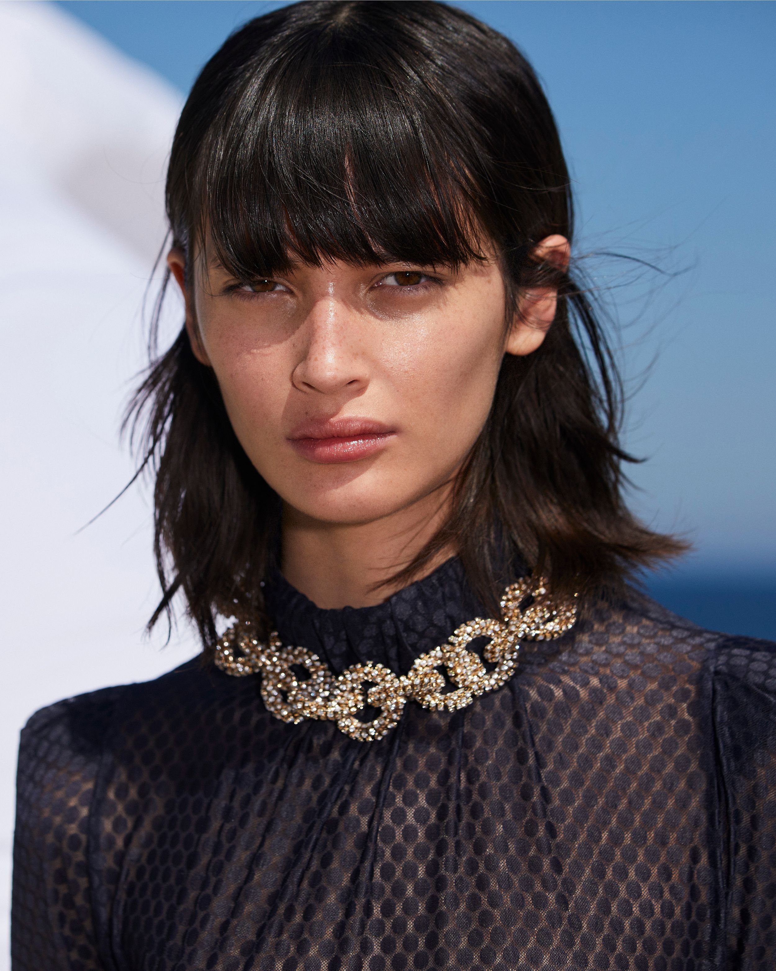 Headshot of model wearing a black sheer blouse and chunky gold diamante necklace